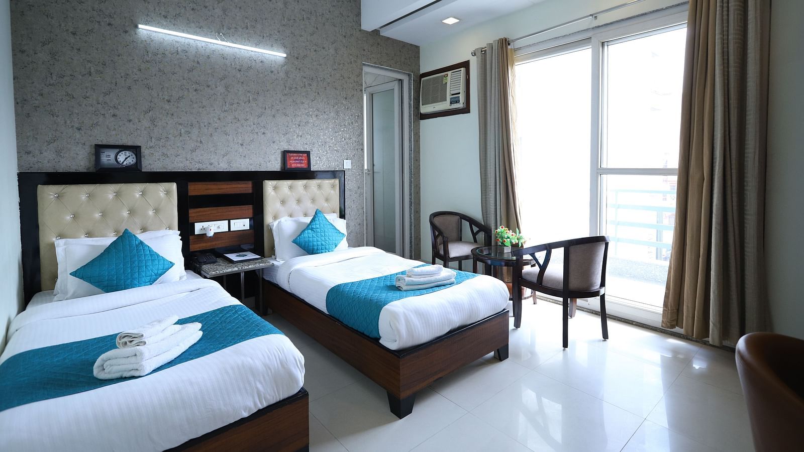A comfortable room contains two single beds with blue accents, dark furniture, and a window with curtains - Asian Suites Cyber Park, Gurgaon