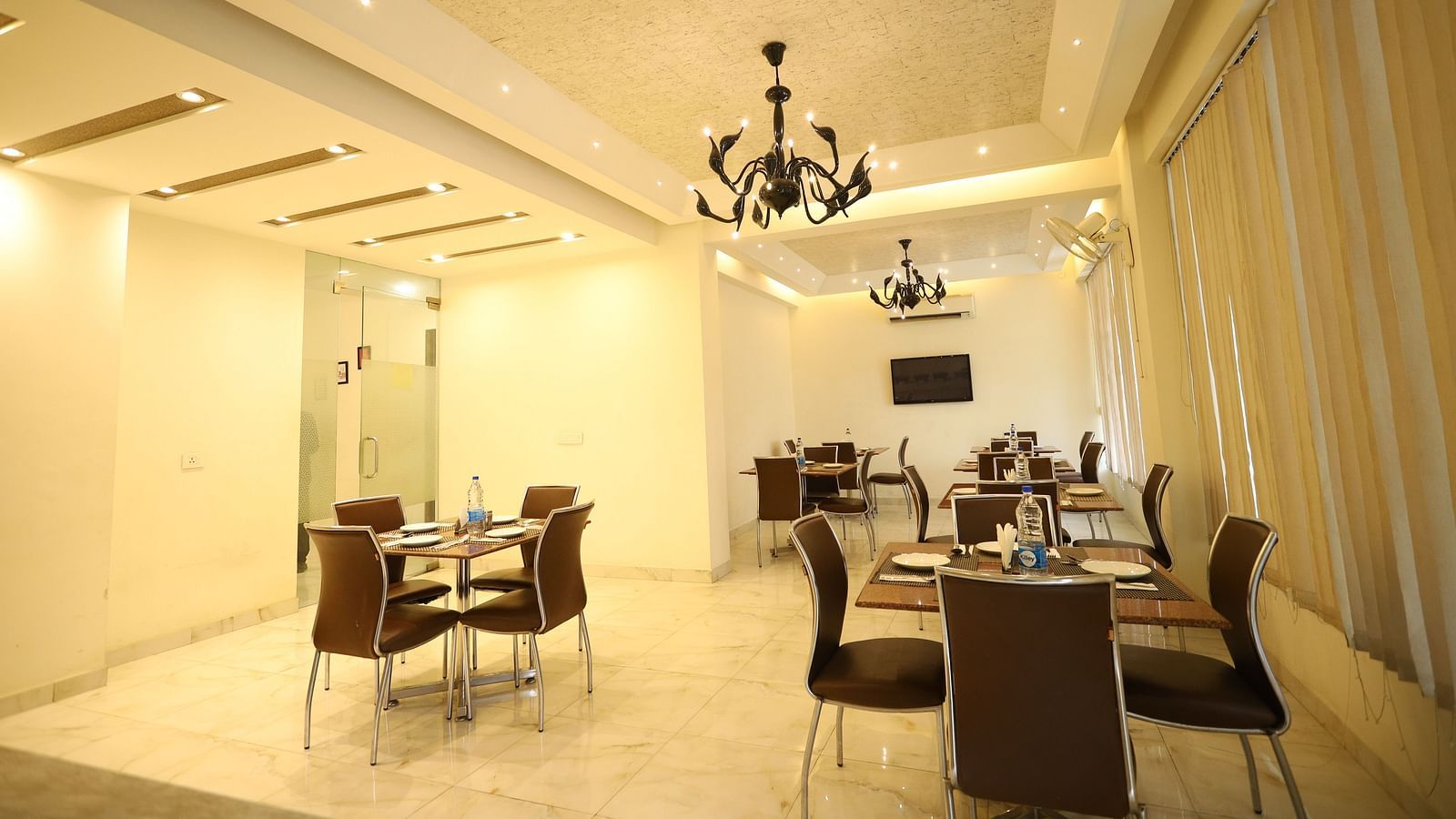 A brightly lit dining area features multiple tables with chairs and modern chandeliers hanging from the ceiling - Asian Suites Cyber Park, Gurgaon
