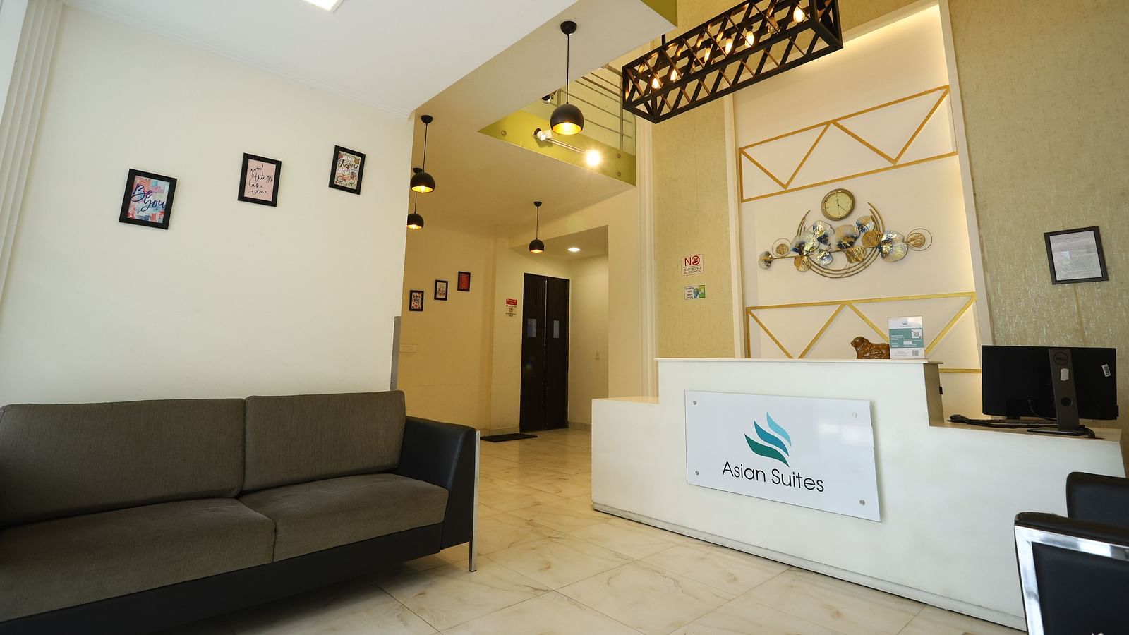 A reception area with a dark sofa and a white counter featuring a logo, illuminated by modern lighting fixtures - Asian Suites Cyber Park, Gurgaon