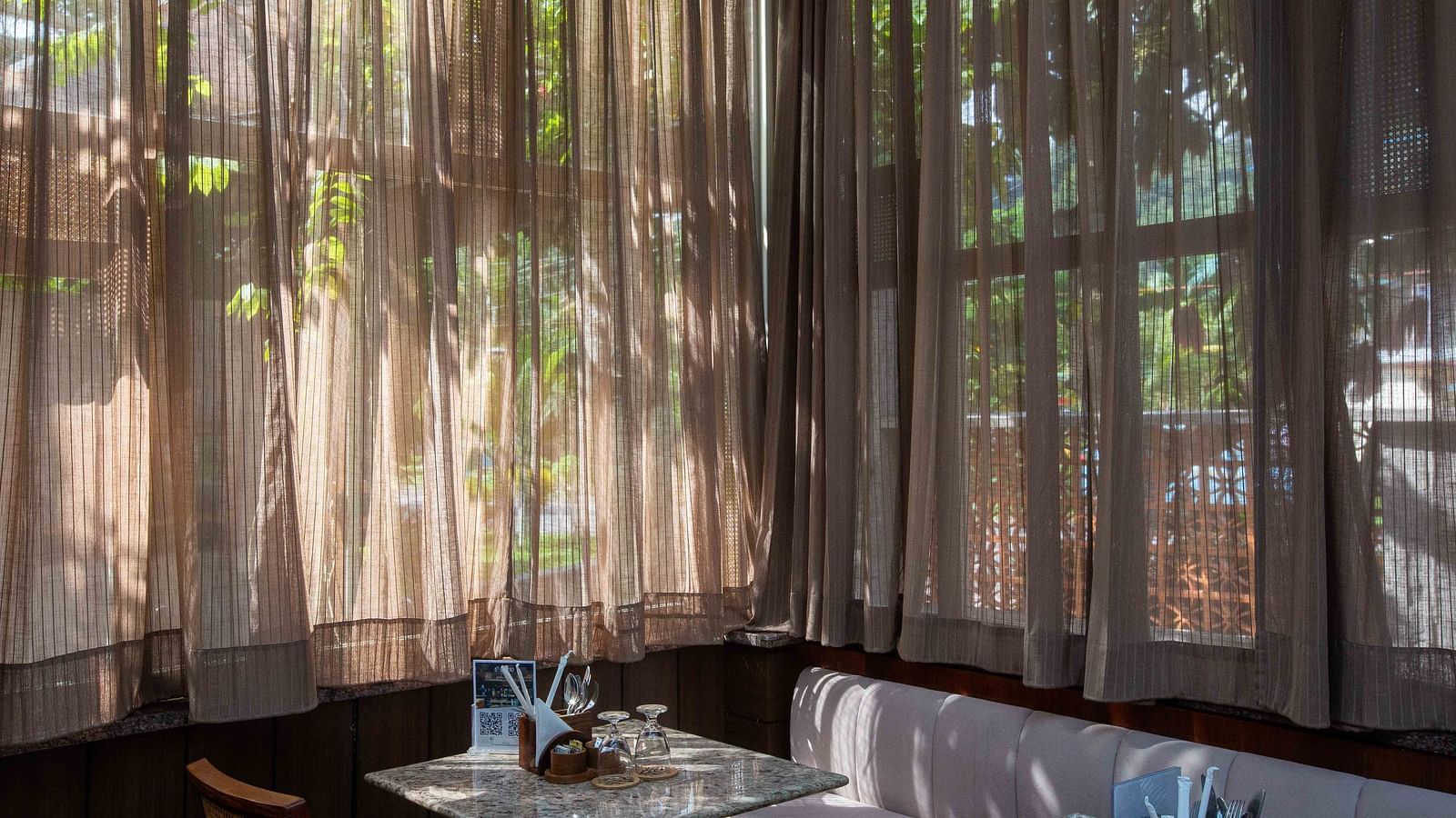An indoor dining area at Ocean Prime Coral Reef Resort & Spa, Havelock with a wide sofa, a marble table, two glasses kept upside down on them and natural light coming through brown transparent curtains - Coral Reef Resort & Spa, Havelock