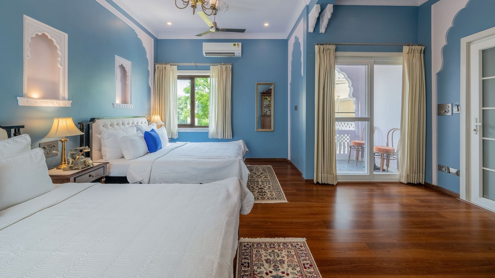 A bright and airy room at Dera Rawatsar, Jaipur, offers two comfortable beds, a balcony with seating, elegant wall details, and wooden flooring.