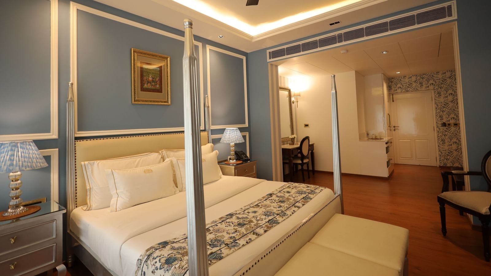 A bedroom with a four-poster bed, blue panelled walls, and a recessed ceiling with warm lighting, leading into an adjacent room with a desk and chair | Dileep Kothi, Jaipur