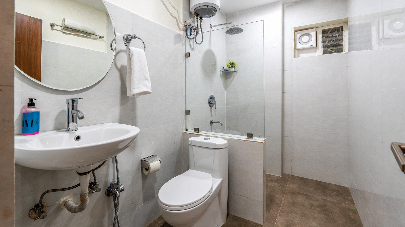 Image of a washroom featuring a circular mirror, wash basin, towel, geyser and a shower area separated by a glass door | Eminent Suites & Apartments, Hi-Tech City 