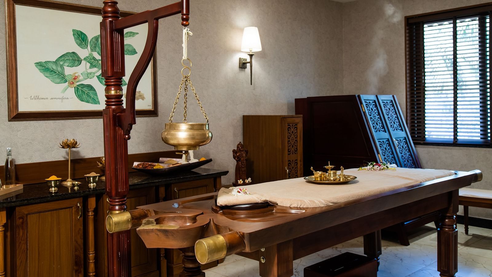 An Ayurvedic treatment room with a wooden massage table and hanging brass pot - Gamyam Retreat