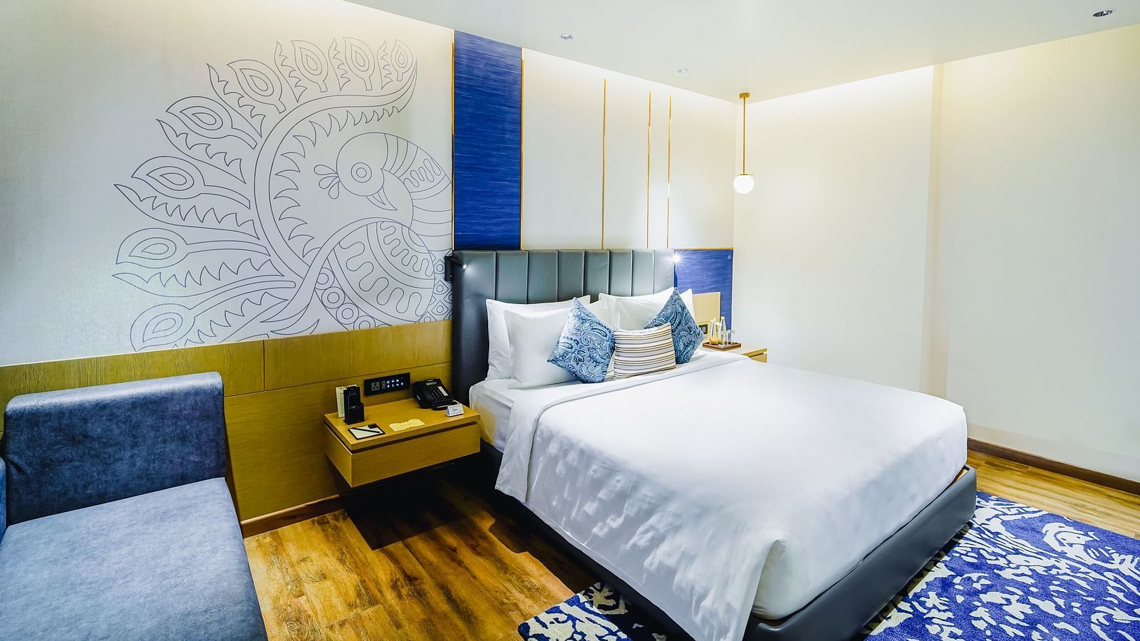 A Deluxe Suite in Madurai with a peacock wall paper, a large bed, a night stand and a blue couch - Grand Madurai by GRT Hotels