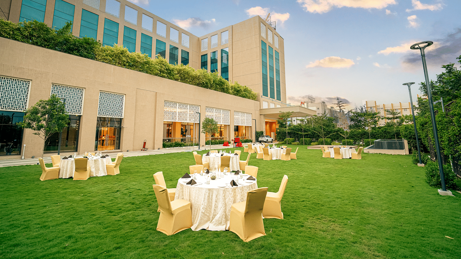 An elegant outdoor event space with round tables set up on a lush green lawn, adjacent to the hotel - Grand Madurai by GRT Hotels