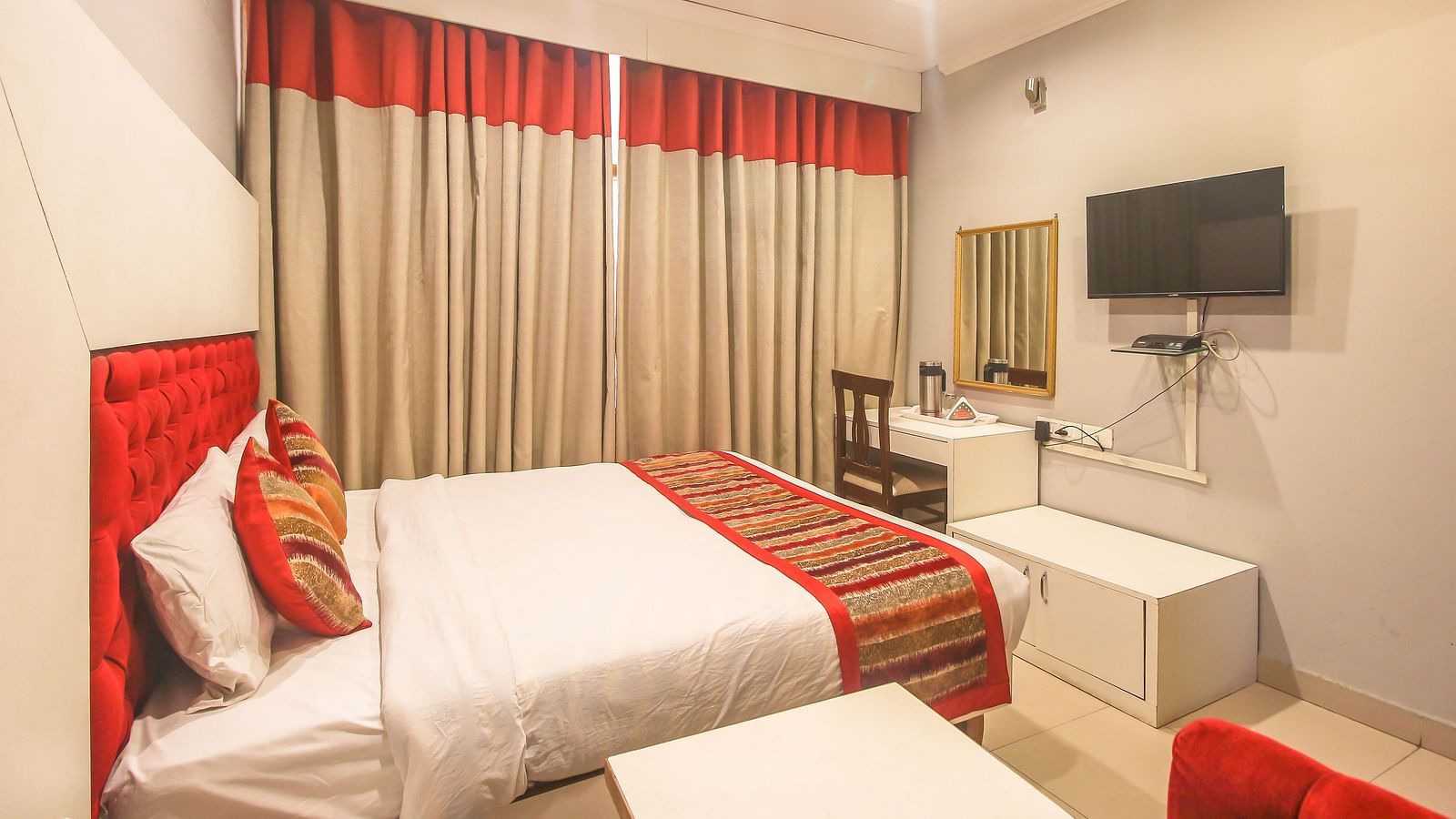A hotel room with a plush bed, layered curtains, a TV, a desk, and a bright red accent chair, illuminated by soft ceiling lights. - Hotel Diamond Plaza, Chandigarh.
