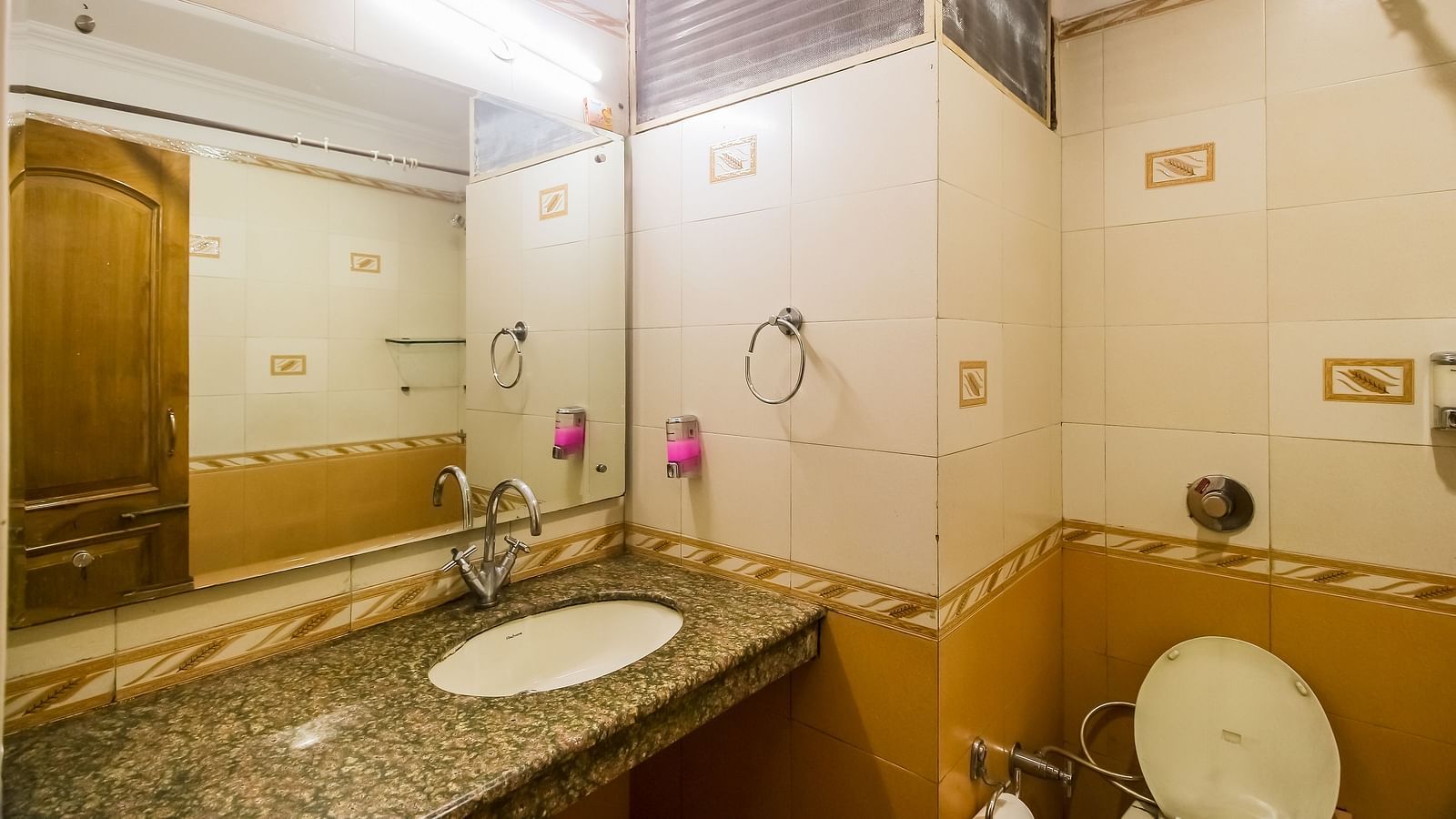 A bathroom with a white oval sink, a granite countertop, a toilet, and a tiled wall with a mirror, illuminated by a light fixture. - Hotel Diamond Plaza, Chandigarh.