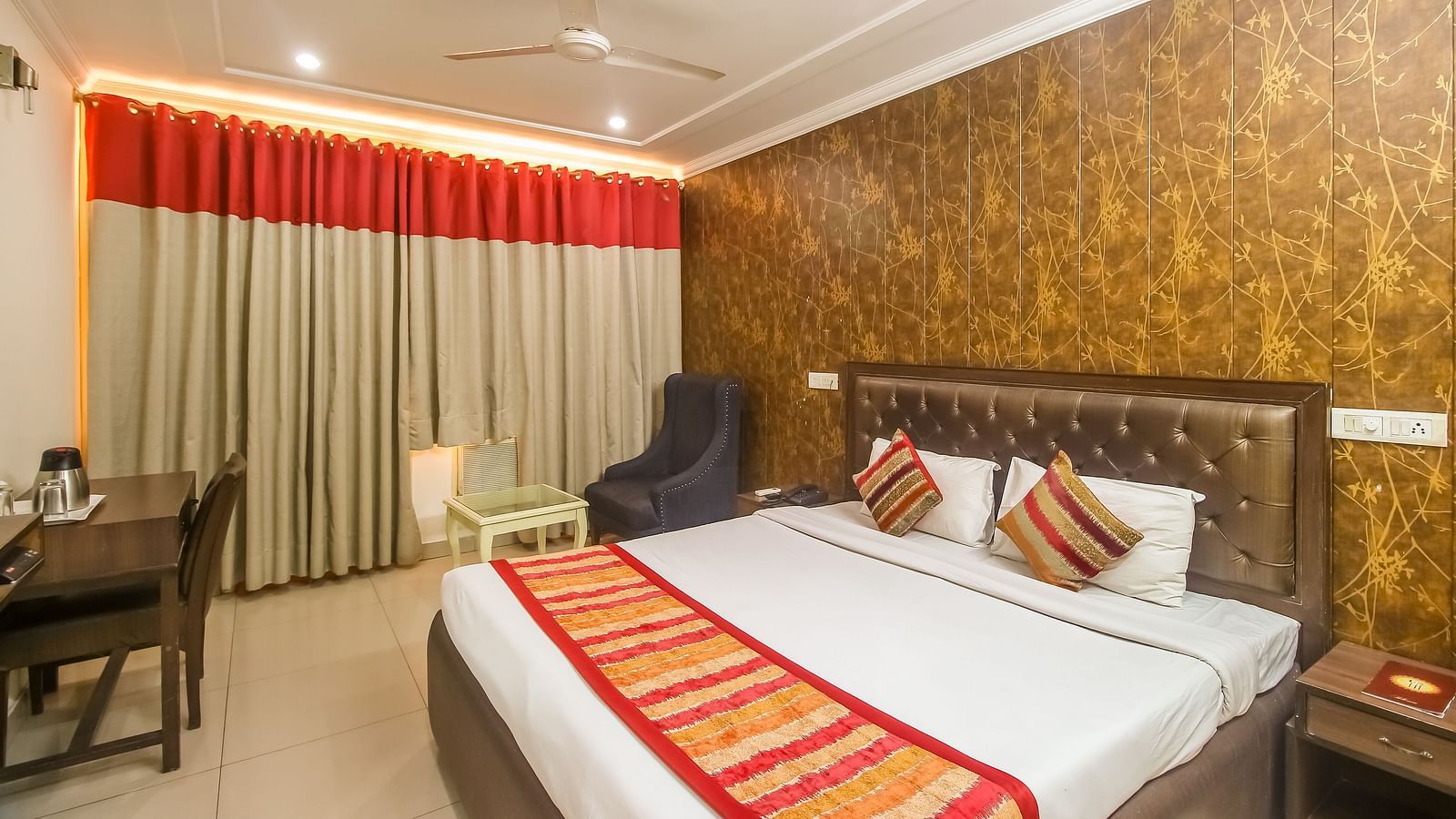 A hotel room with a textured accent wall, a plush bed with decorative pillows and a runner, a dark armchair, and a desk, illuminated by warm lighting. - Hotel Diamond Plaza, Chandigarh.