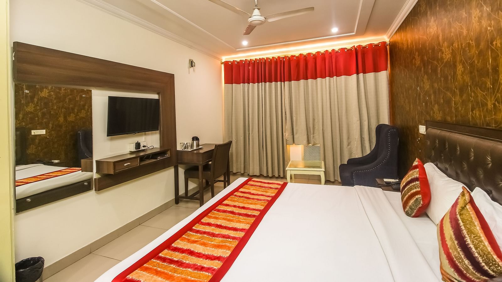 A hotel room with a plush bed featuring a patterned runner, a TV, a desk, a dark armchair, and a textured accent wall, lit by soft lighting. - Hotel Diamond Plaza, Chandigarh.