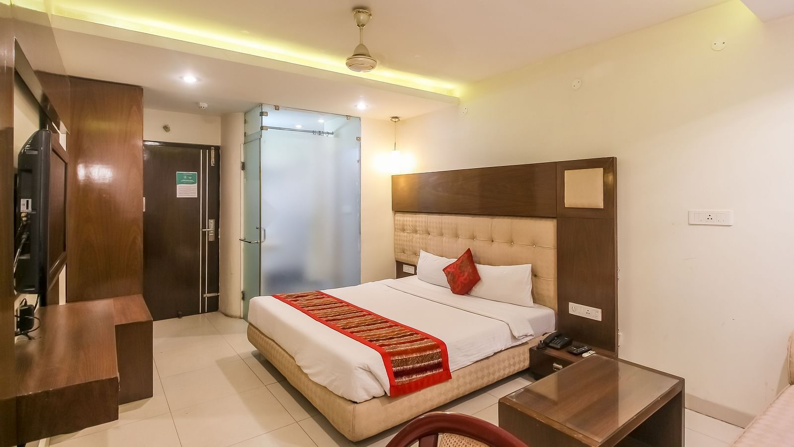 A hotel room with a neatly made bed, wooden furniture, and a glass-panelled bathroom. - Hotel Diamond Plaza, Chandigarh.