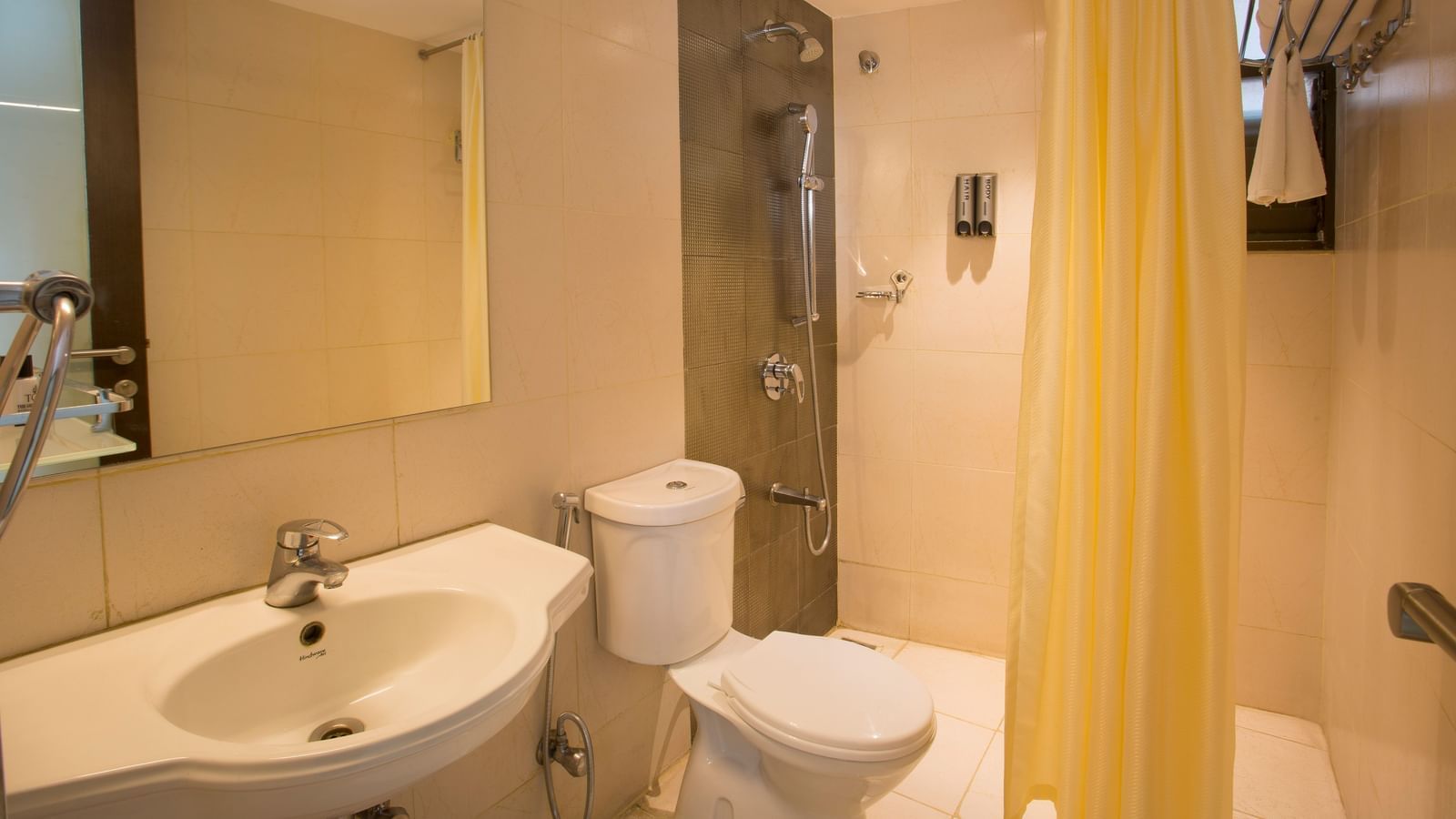 Another view of a hotel bathroom with a beige tiled shower area, white toilet, sink and a mirror, with a yellow shower curtain - Hotel Langford Keys by TGI, Shanti Nagar
