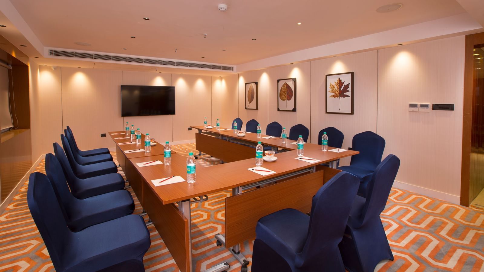 A formal boardroom with blue chairs around a long table, a flat-screen TV on the wall and framed leaf prints - Hotel Southend by TGI - Bommasandra, Bangalore