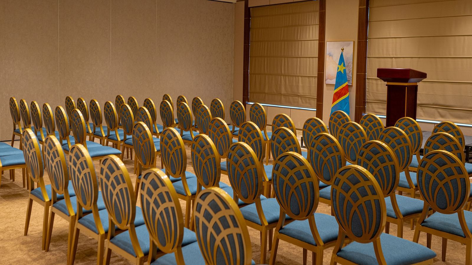 Rows of empty chairs at KertelSuites are arranged facing a podium in a well-lit conference room with beige walls and large windows covered by blinds