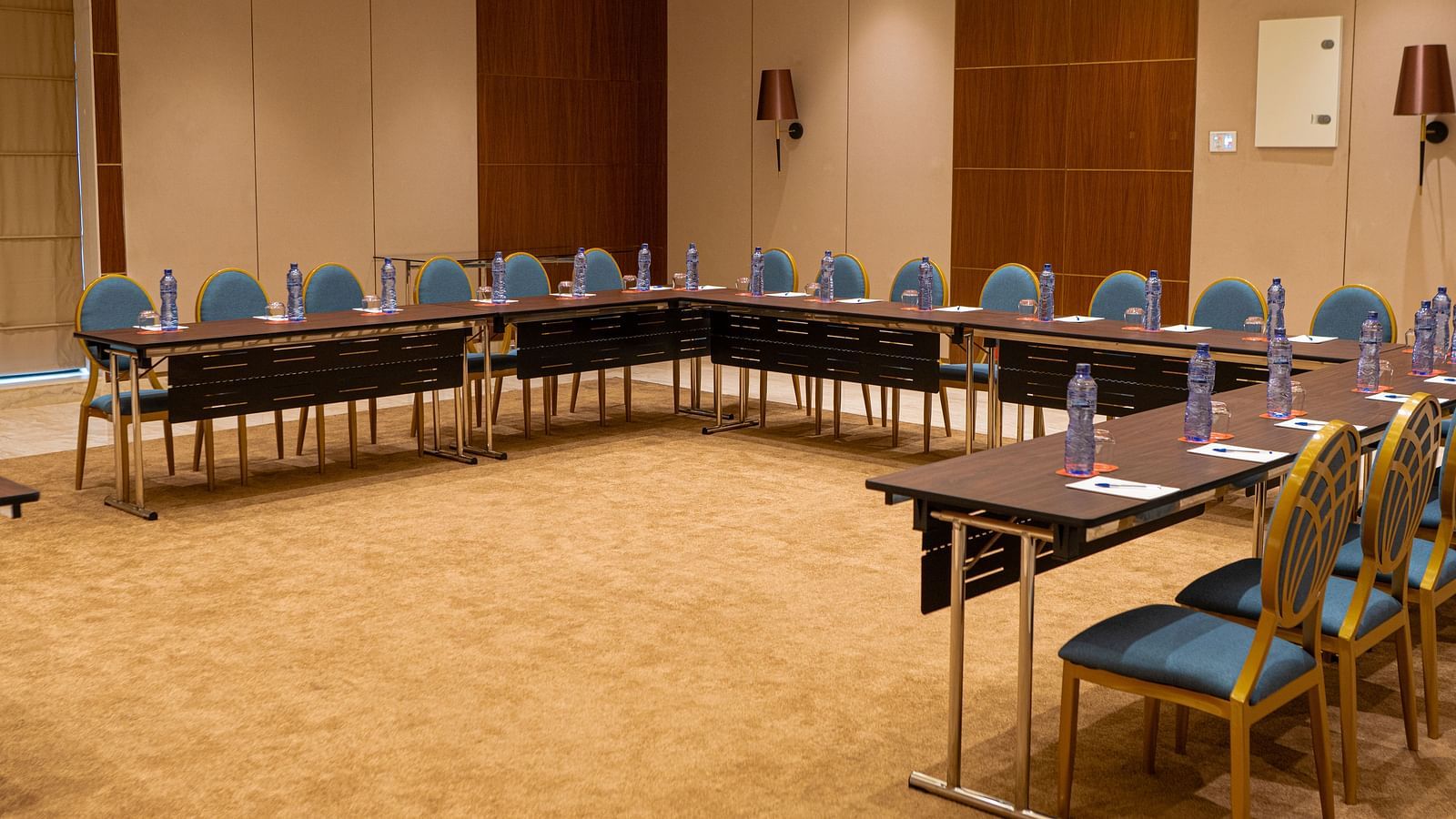 A conference room at KertelSuites with a U-shaped arrangement of tables and chairs, each table set with bottled water and notepads, in a well-lit, carpeted space