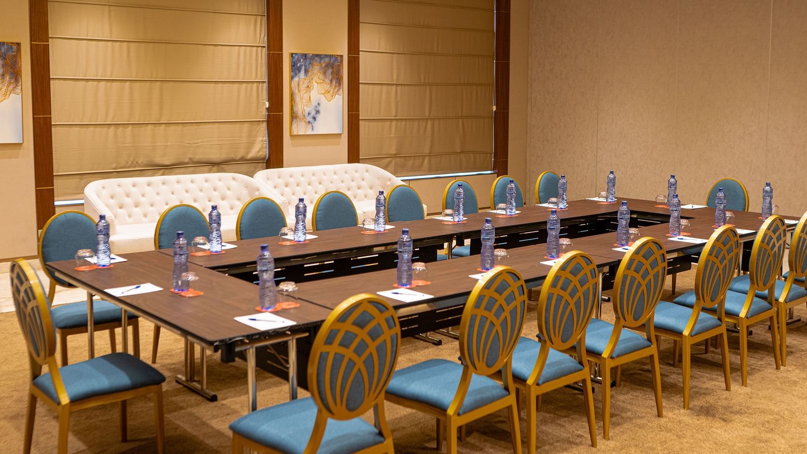 A conference room at KertelSuites with a large U-shaped table setup, chairs, bottled water, notepads, and pens at each seat. The room has beige walls, wood accents, and framed artwork.