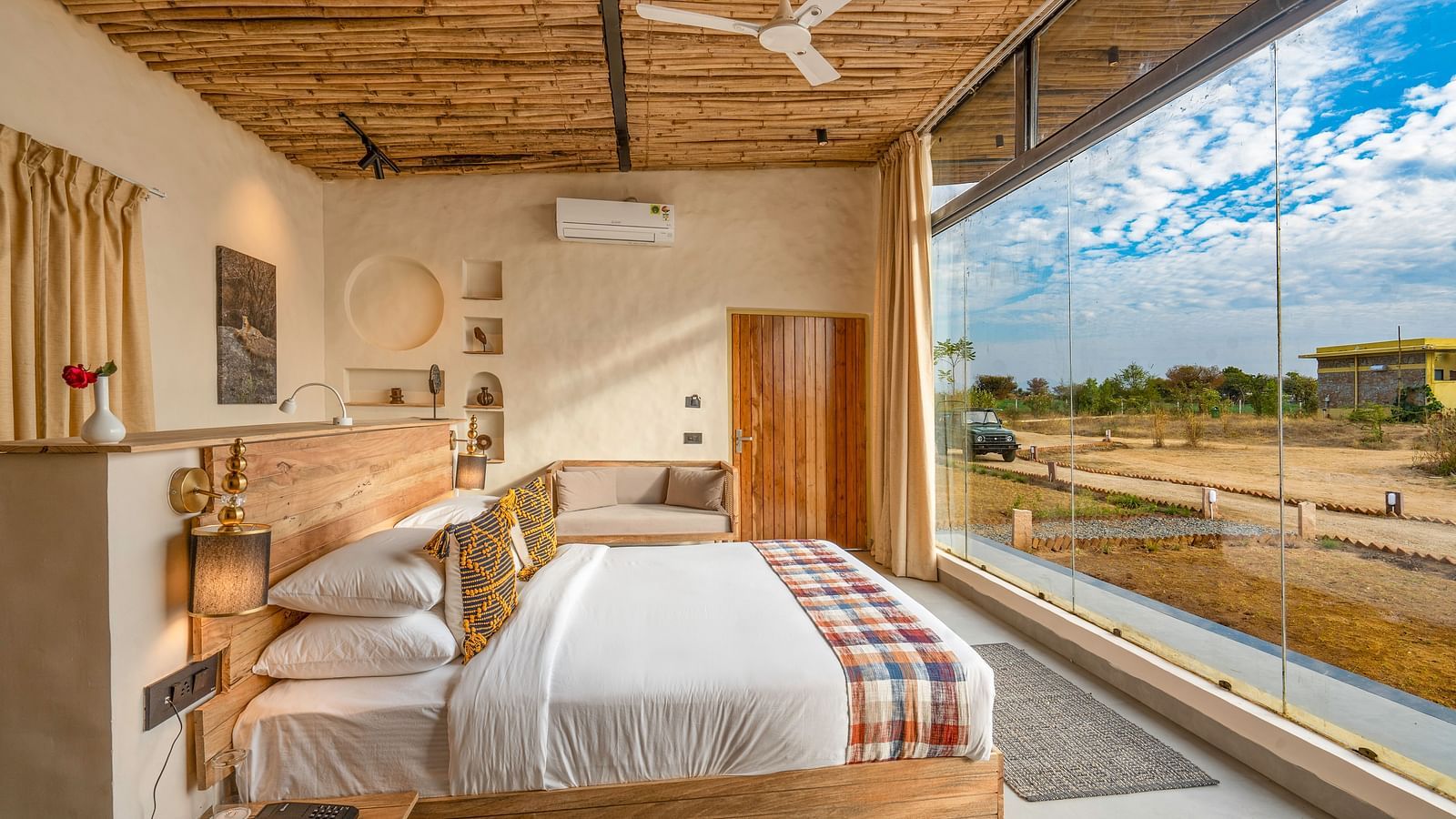 An image of a wooden furnished bedroom featuring a double size bed, a bedside table with lamp and socket, a sofa, some decorative elements in the room, and a large window from where outdoor scenery can be enjoyed - Kurja Jawai