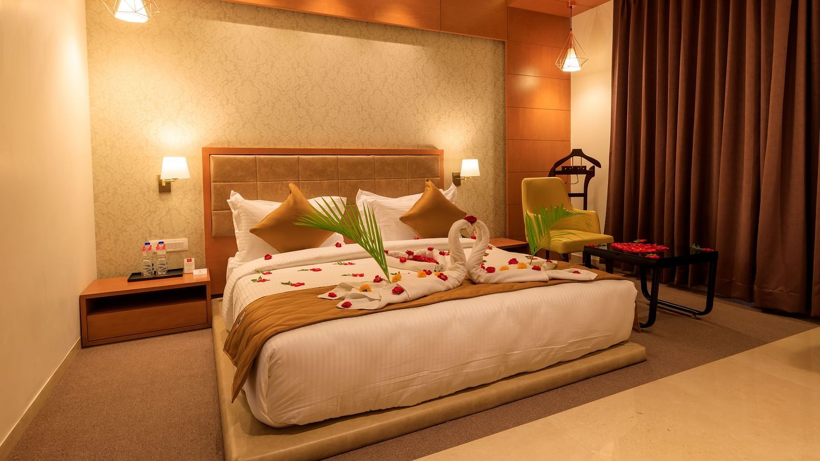 A warmly lit hotel room at Lords Eco Inn Apple Gate features a decorated double bed, bedside tables, a chair, and brown curtains.