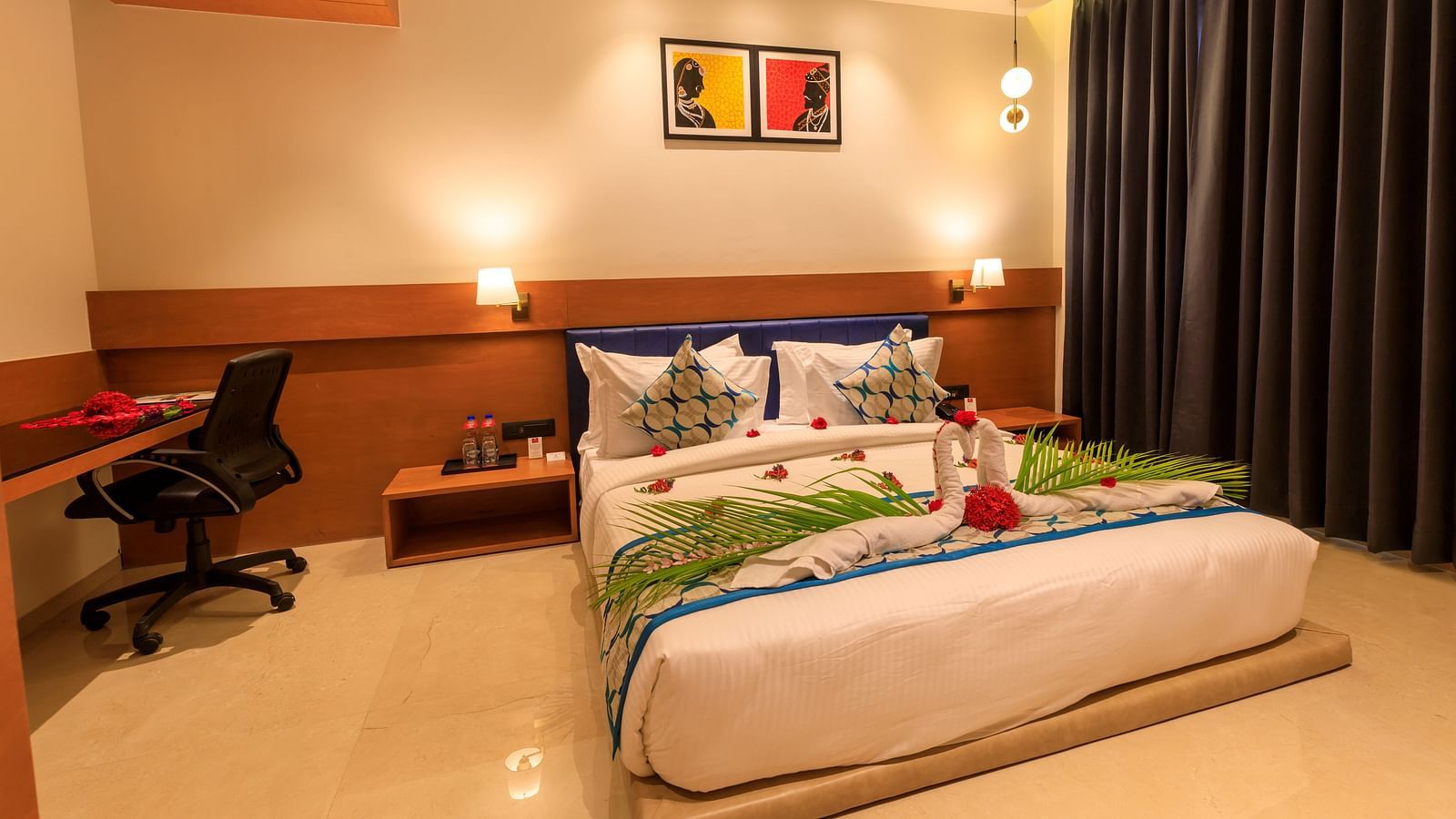 A warmly lit hotel room at Lords Eco Inn Apple Gate features a decorated double bed, bedside tables, a desk with a chair, and dark curtains.