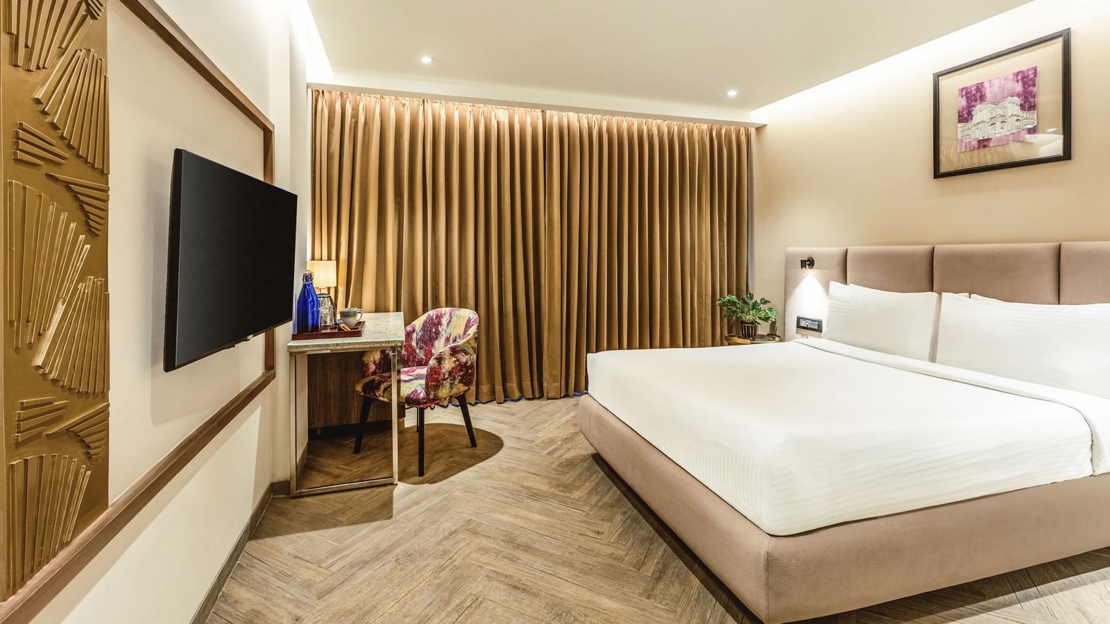 A wide-angle view of a room with a large bed and a television at Mango Hill Central, Chennai. A desk and chair are visible on the right.
