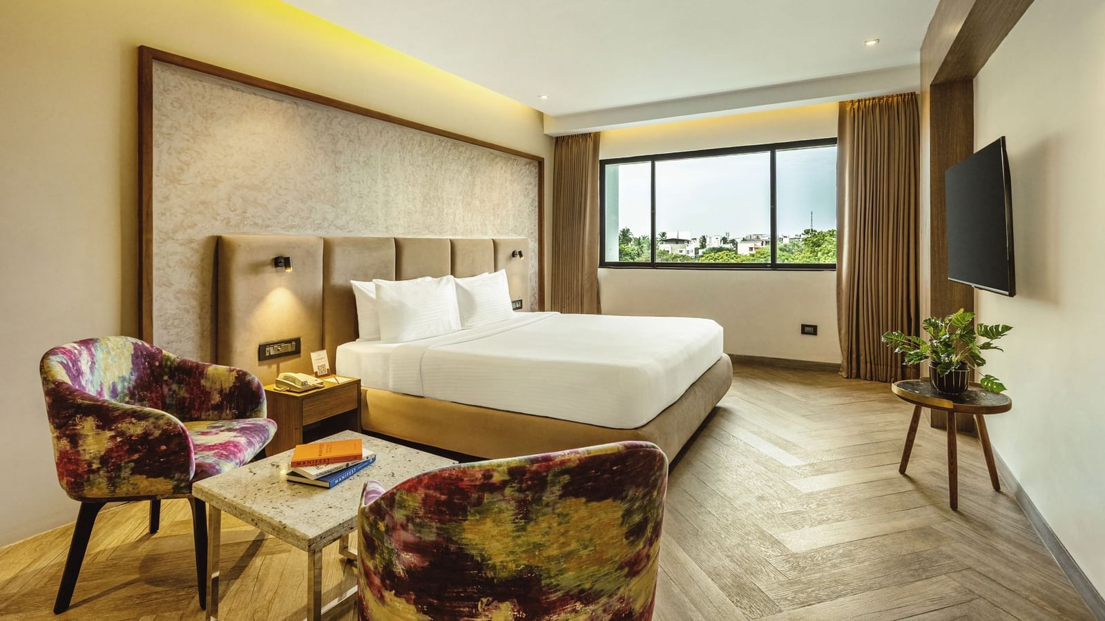 A wide-angle view of a room with a large bed at Mango Hill Central, Chennai. There are a chair and a small table in the foreground.