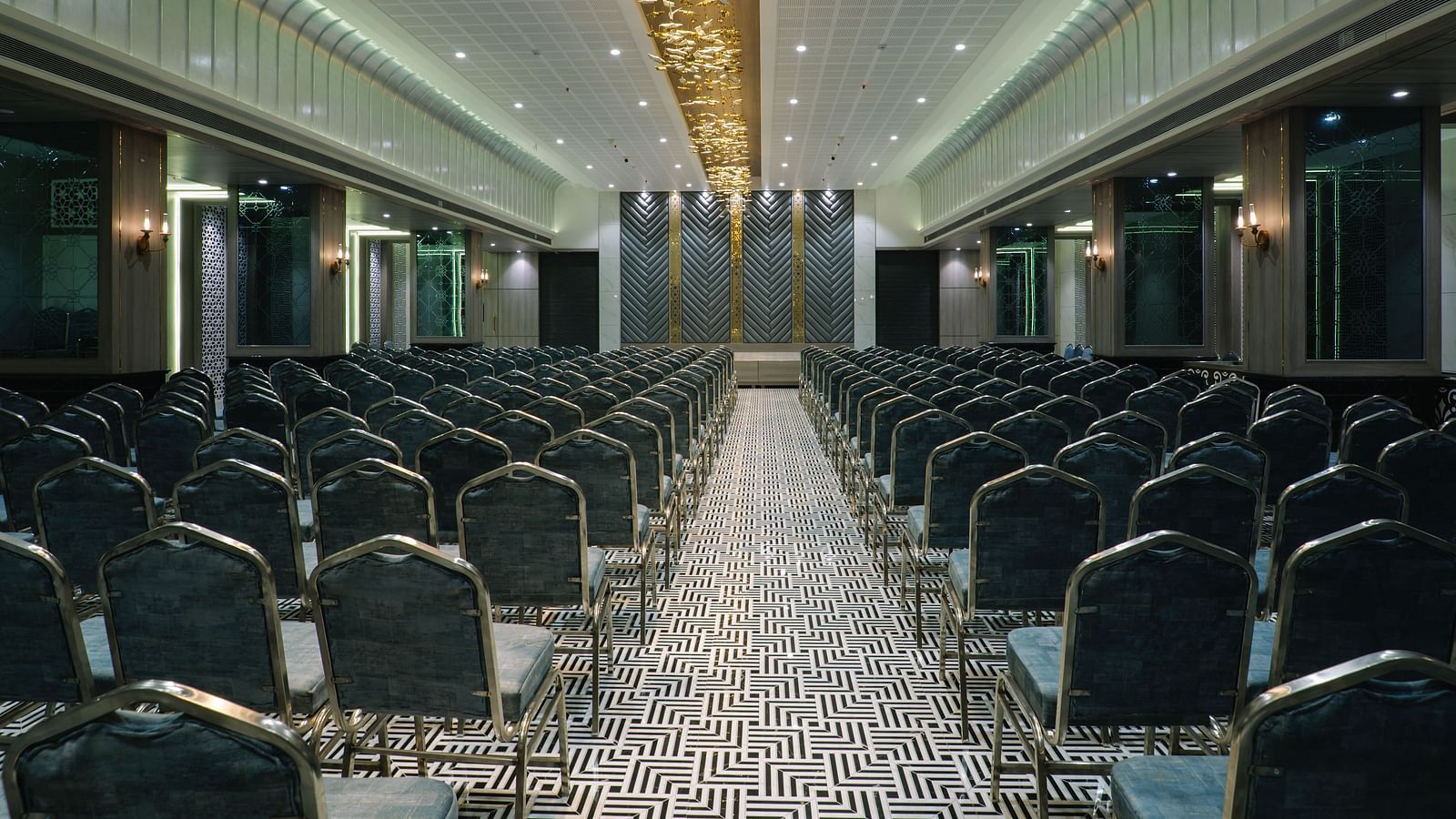 A large room is set up with rows of dark chairs facing a stage or presentation area - Melior Inn