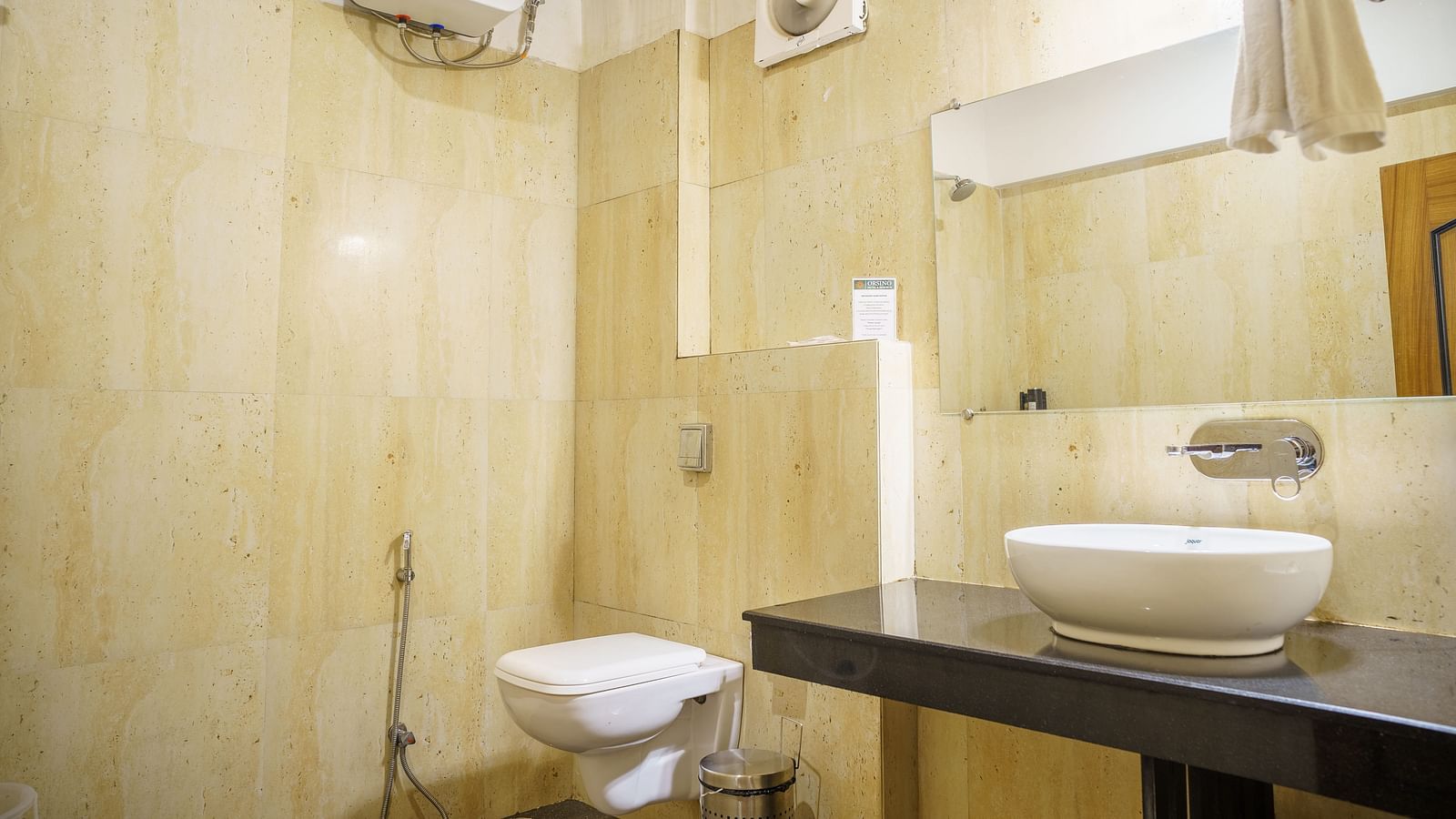 A bathroom at Orsino Spa Resort with a white basin on a dark countertop, a toilet, and light-coloured tiled walls.