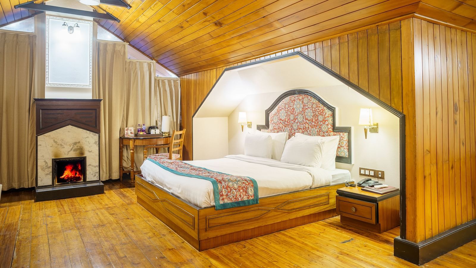 A room at Orsino Spa Resort with a pitched wooden ceiling, a bed with a patterned throw, and a small fireplace.
