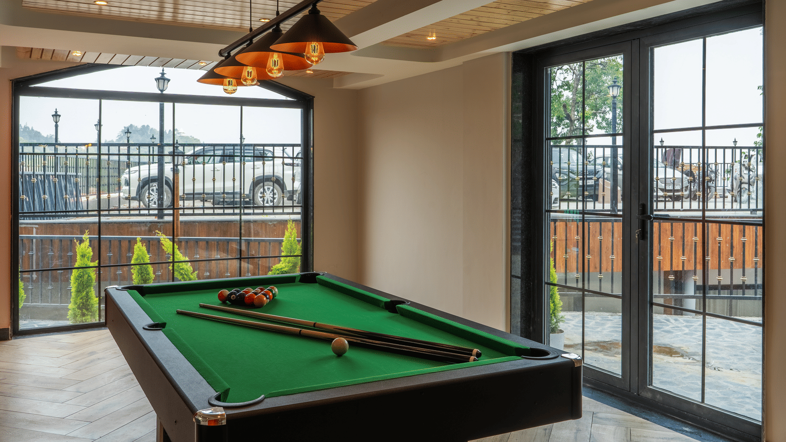 A green billiard table with cues, inside a room with large glass windows and hanging lights at Perfectstayz Luxe Mall Road.
