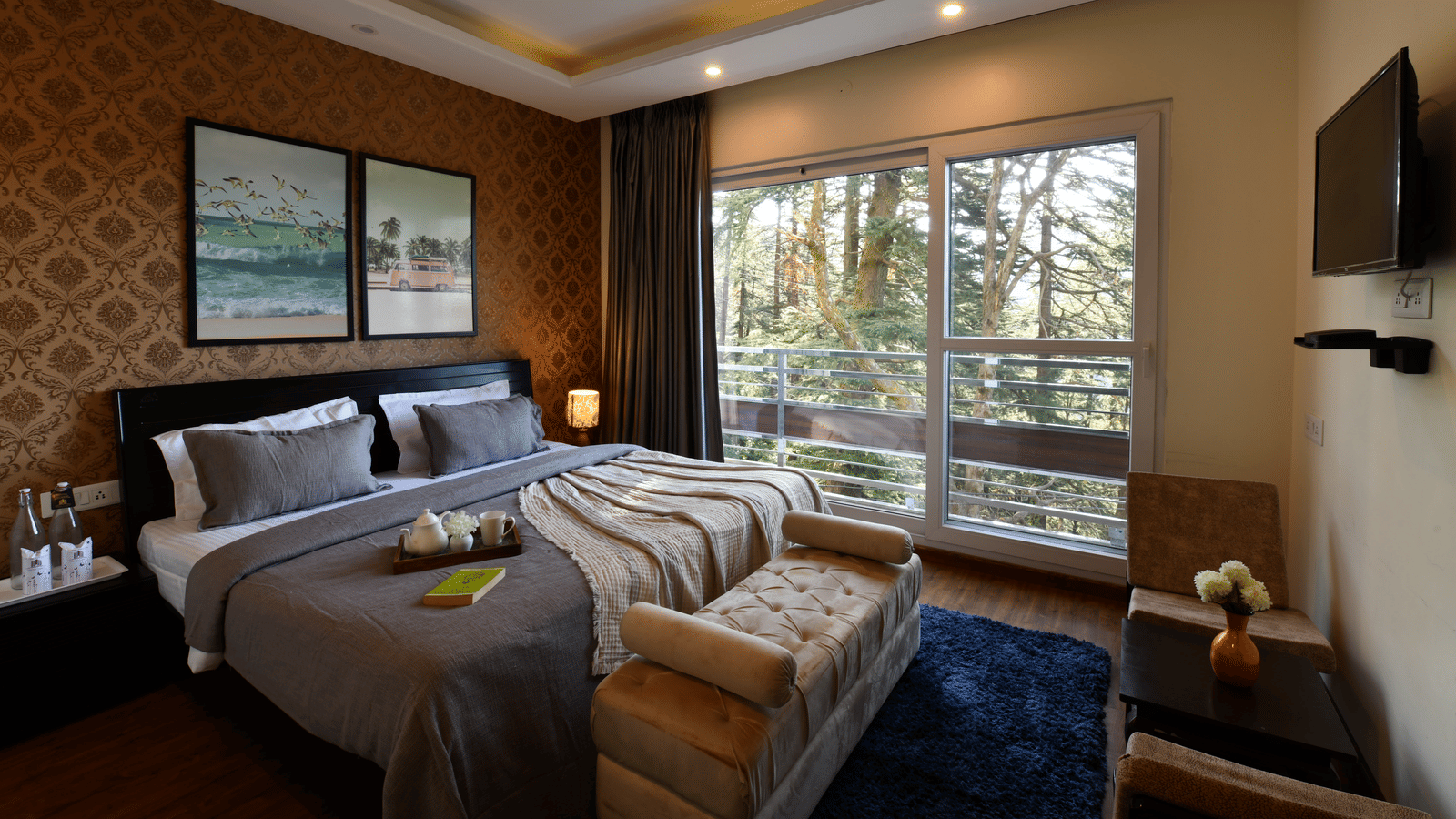 A bedroom at Perfectstayz Value Shimla (Namah Retreat) with a king size bed, a daybed, wall paintings, a television, a tray with cups and kettles, a table, 2 chairs, and a sliding door leading to a balcony.