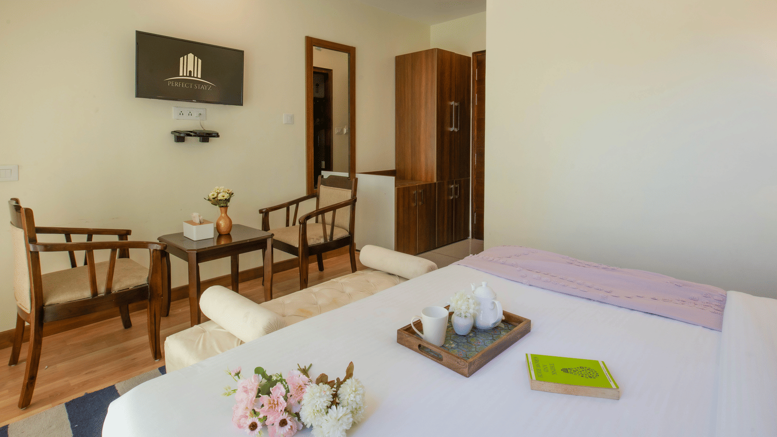A bedroom at Perfectstayz Value Shimla (Namah Retreat) with a king size bed with a tray of cups and a kettle, 2 wooden chairs, a small table, wall art, a mirror, a cabinet, and a cupboard.