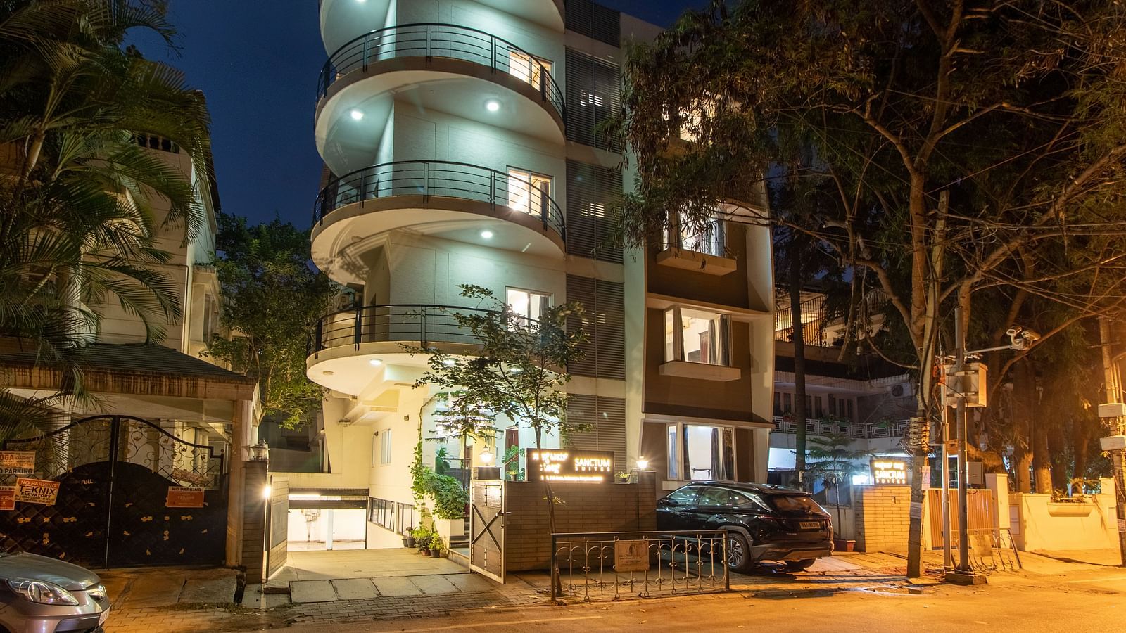 An exterior view of Sanctum Suites Indiranagar, Bangalore showcasing the cylindrical building illuminated at night along with greenery around.