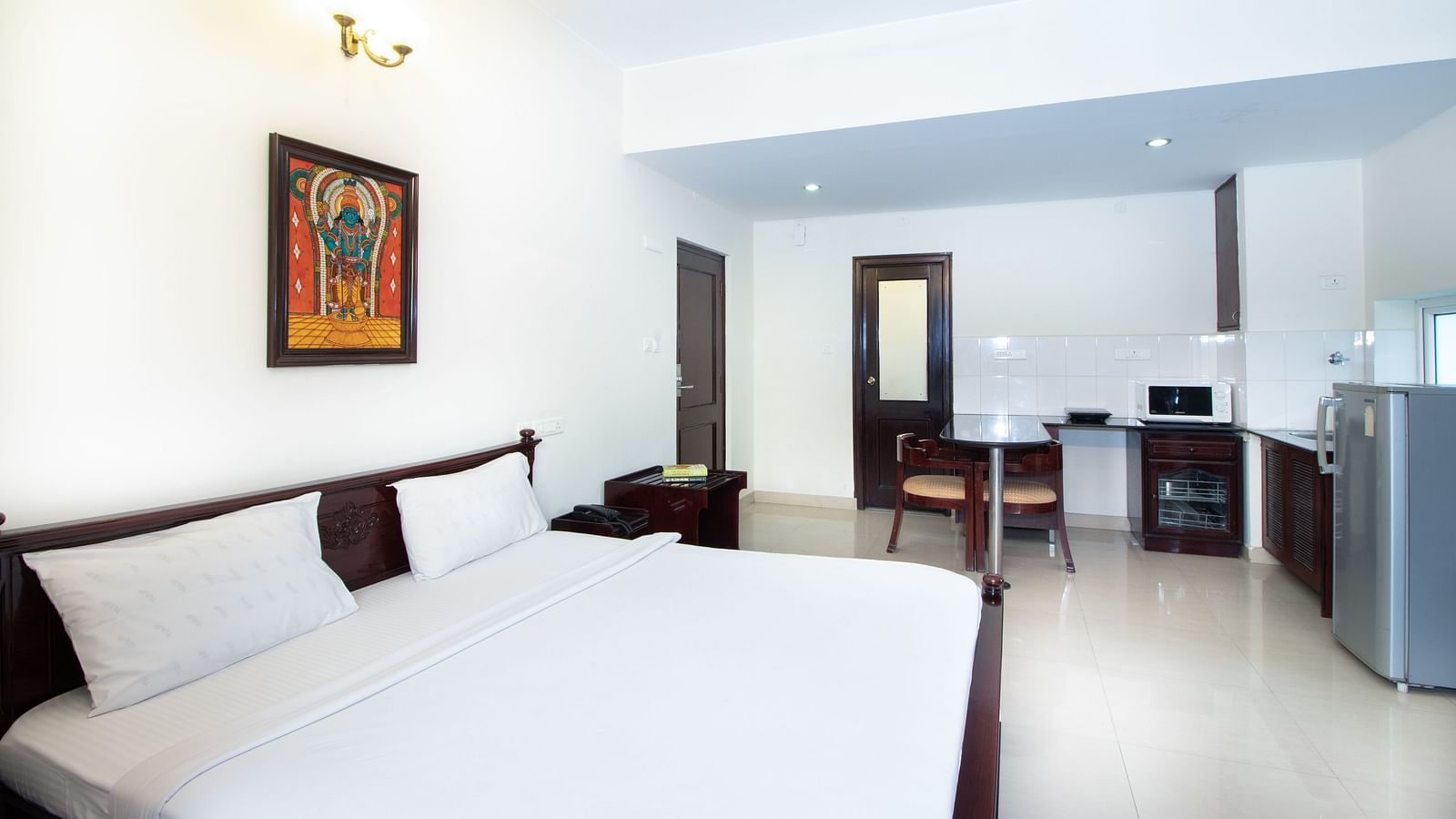 A room with a double bed featuring white linens, a ceiling fan, a framed picture on the wall, a dining counter, a refrigerator, and the kitchenette area at Sanctum Suites Indiranagar, Bangalore.