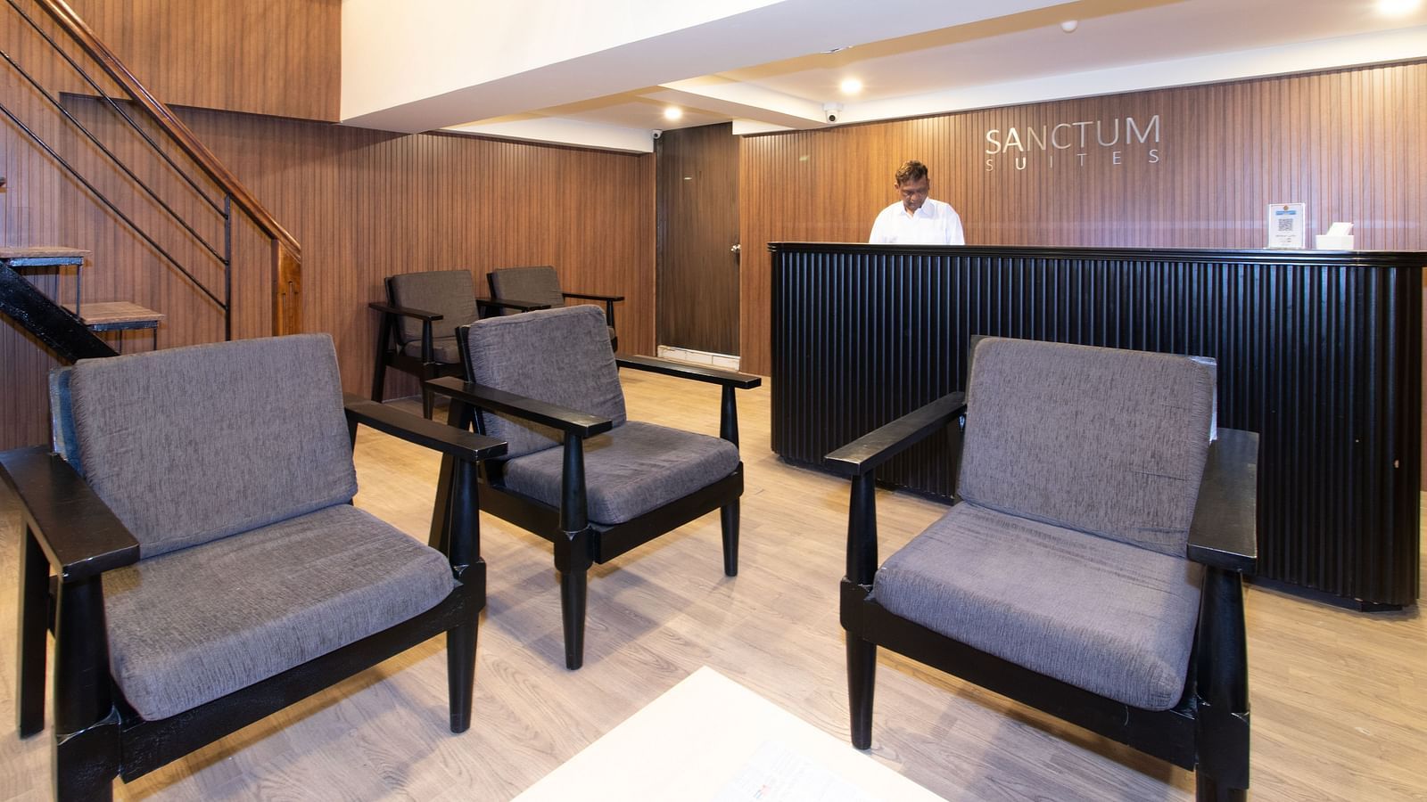 Reception area with grey armchairs, a reception desk, and a person in the background - Sanctum Suites Richmond Road, Bangalore