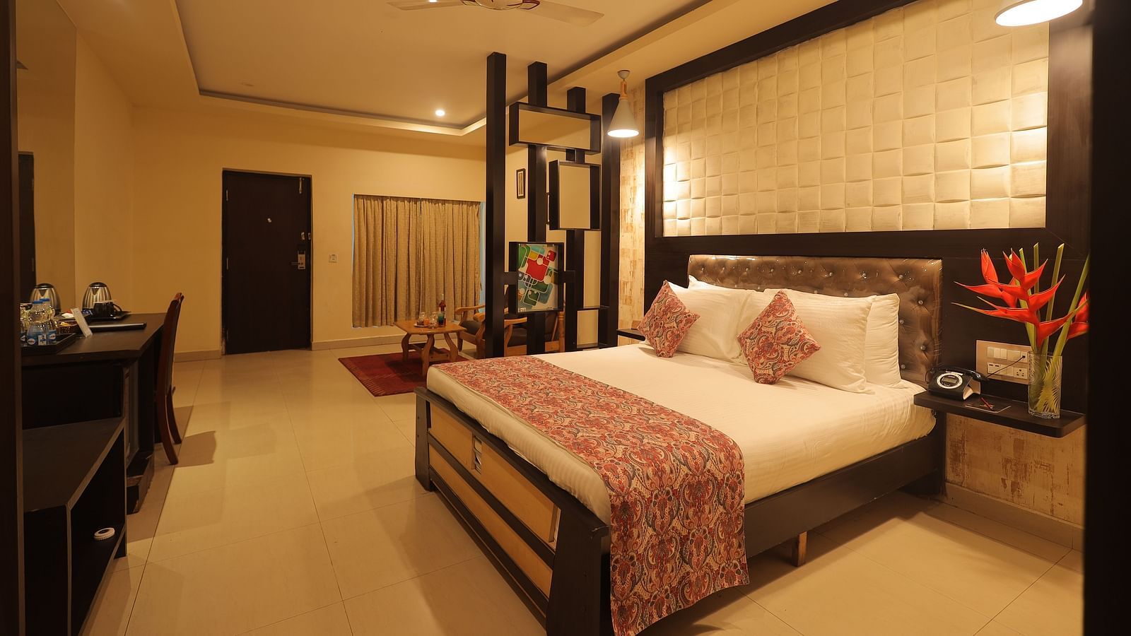 An image of a bedroom with bedside table, a set of table and chairs, flat TV screen, and AC- Sathyam Grand Resort