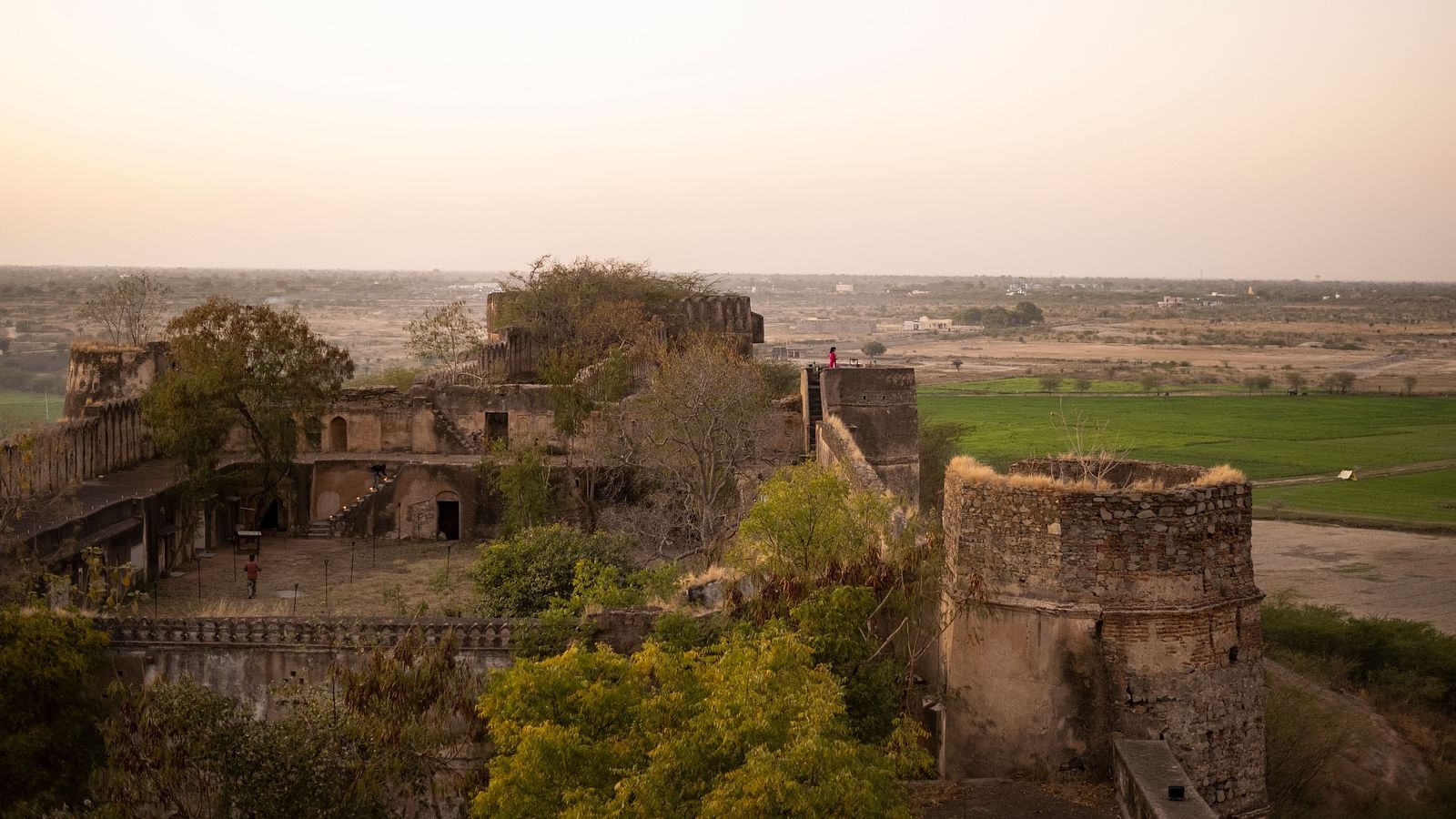 An aerial view of a historic fort with stone walls and towers, overlooking a landscape with trees and distant fields under a hazy sky- Shahpura Bagh.