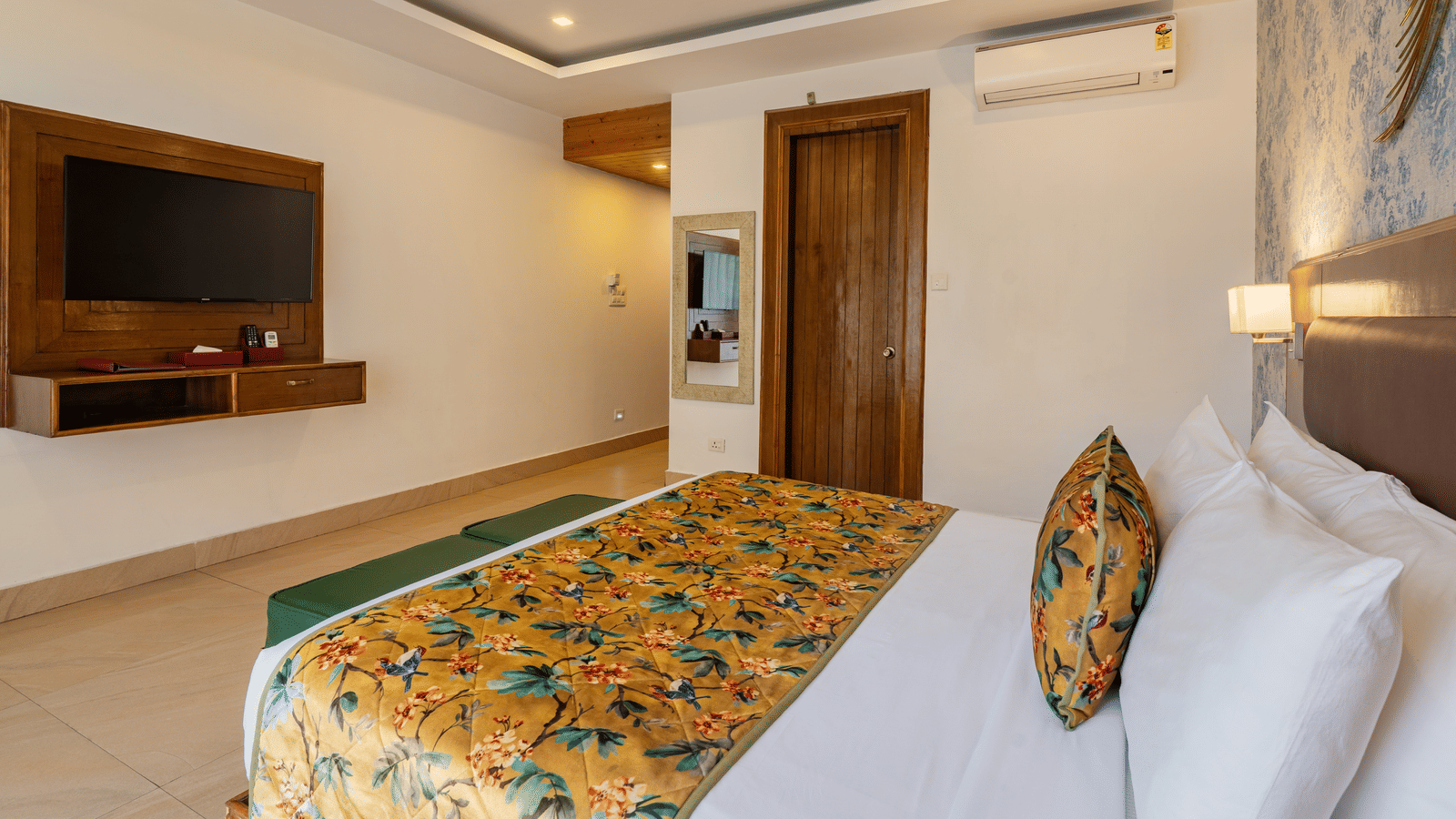 A room with an attached bathroom, featuring a bed, a television unit, a small wall mirror, a decorative ceiling and tiled floor | Summit by the Ganges Beach Resort & Spa, Rishikesh