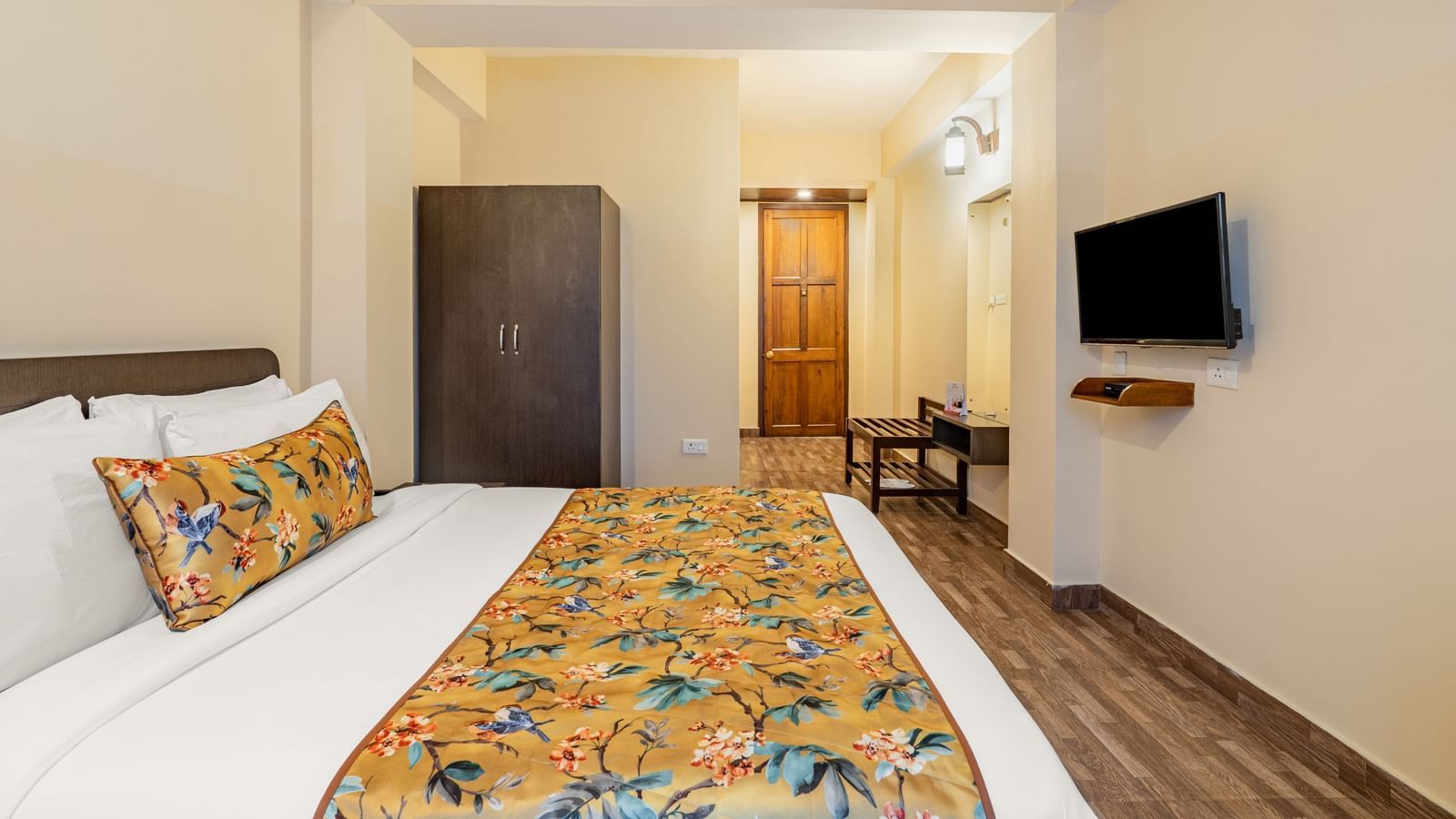 Spacious hotel room with a double bed, floral yellow bedding, wardrobe, wall-mounted TV, wooden flooring, and seating area near the entrance - Summit Namnang Courtyard & Spa, Gangtok