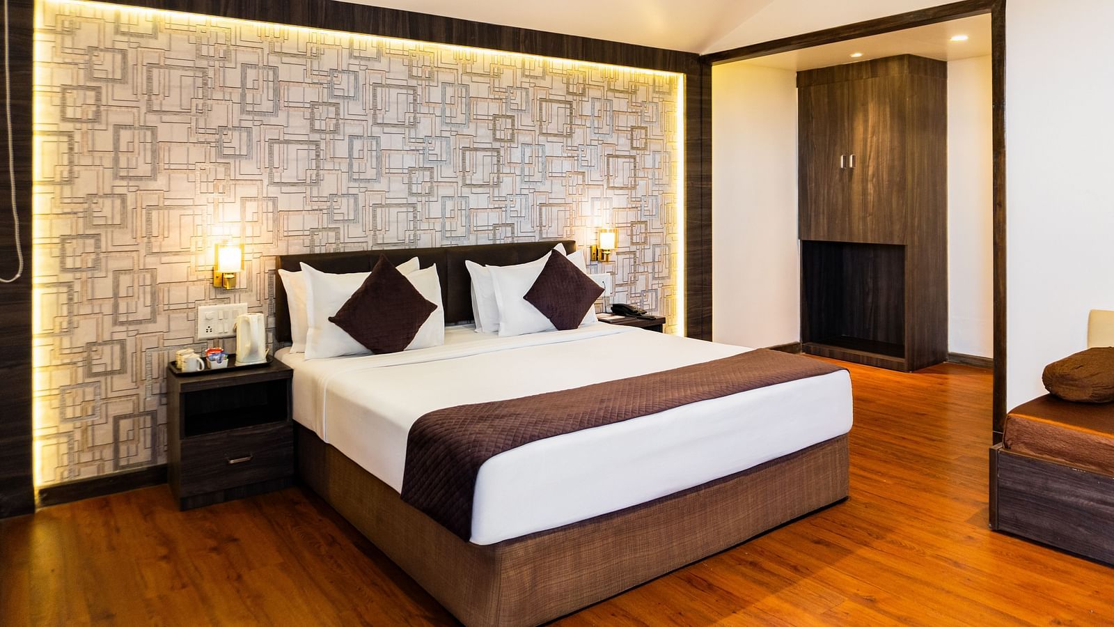 Modern hotel room with a king size bed, warm wooden flooring, geometric accent wall with soft backlighting, bedside tables, and cosy ambient lighting - The Grand Legacy Resort & Spa
