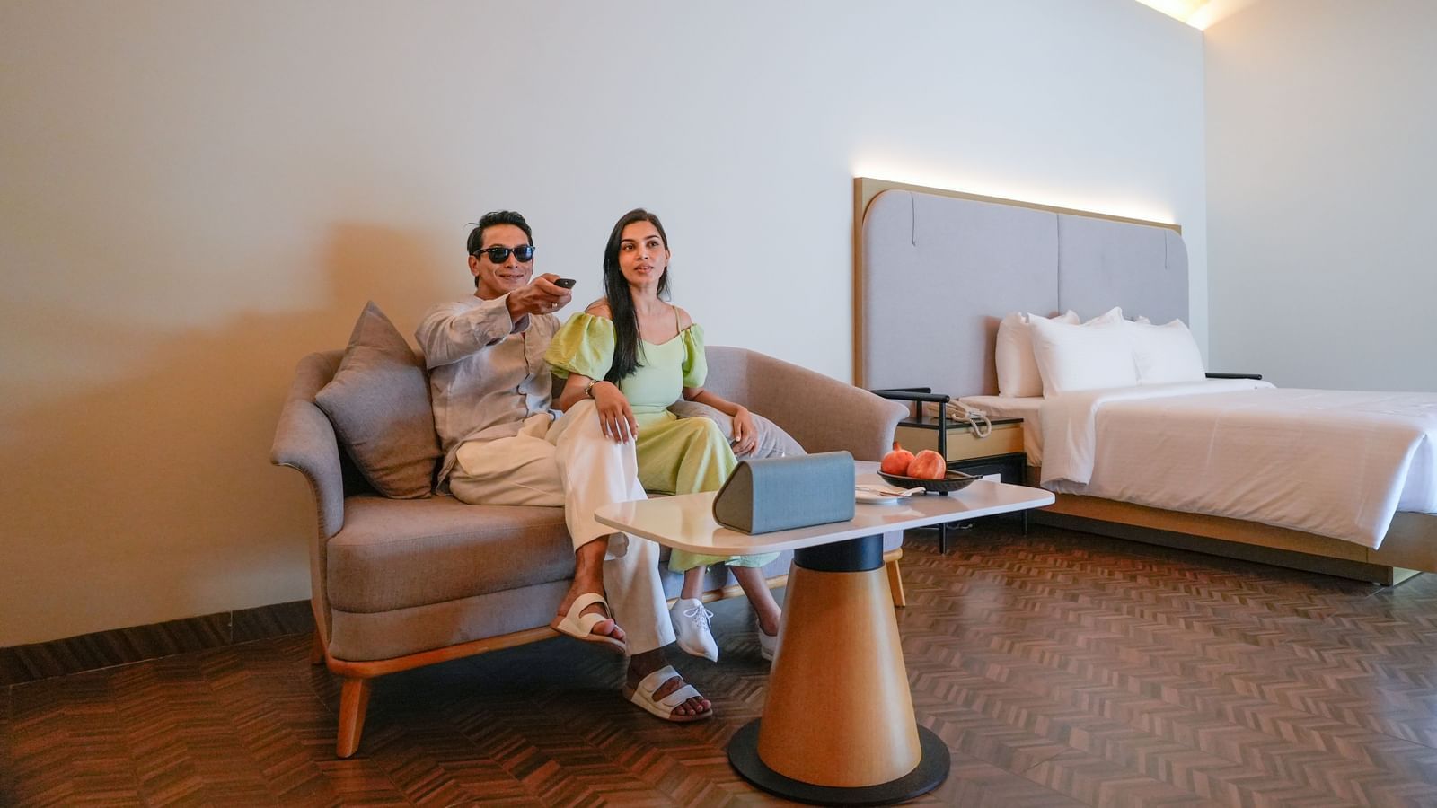 A couple sits on a modern sofa in a stylish hotel room at Exoctica Pool Villa, watching TV. The room features a plush bed, soft lighting, and a coffee table with fruit - The Shore Trishvam, Pondicherry