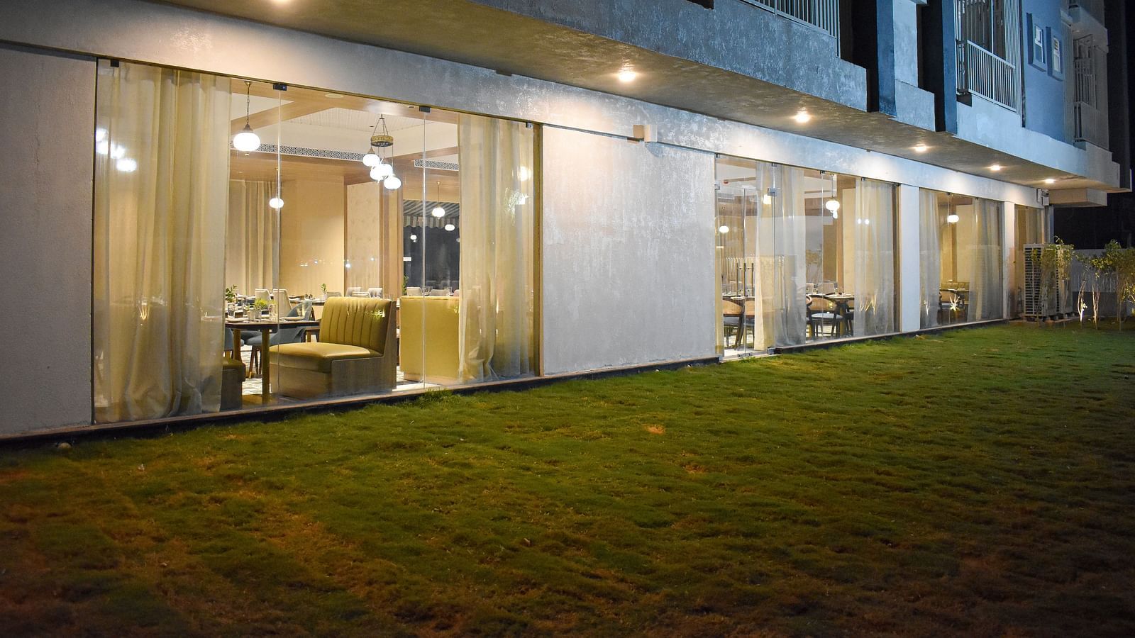 Exterior view of a building with a glass-walled restaurant, sheer curtains, hanging lights, and a grassy area in front - Urban Suites by BluSalzz, Jaipur
