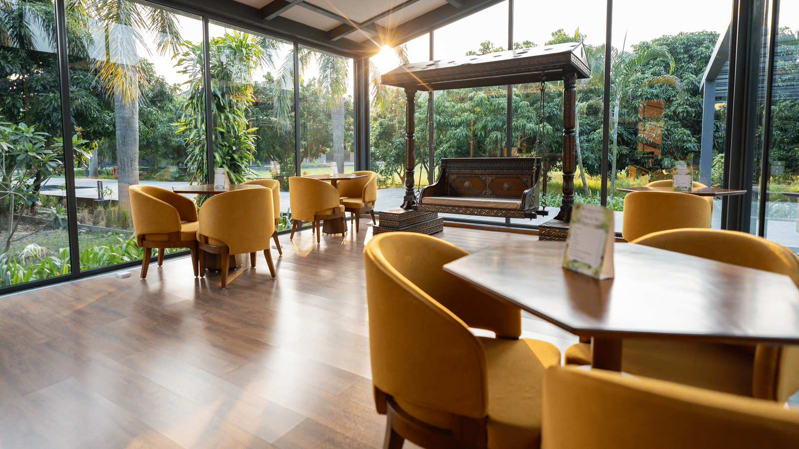 A bright glass-enclosed seating area at Wyndham Garden featuring round tables with upholstered chairs, a wooden swing bench, and views of lush outdoor landscaping.