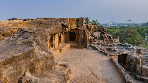 Khandagiri caves, an ancient rock-cut complex on a hill, showing carved openings and pillars, overlooking a distant city skyline and forest.
