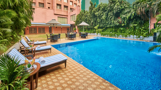 An outdoor swimming pool during the day with blue water, lounge chairs, and a multi-story building in the background | HHI Varanasi