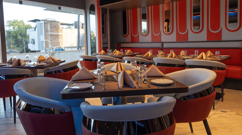A restaurant interior with individual tables set with cutlery, featuring chairs, booth seating with red accents, and large windows | Pride Elite Chhatrapati Sambhajinagar