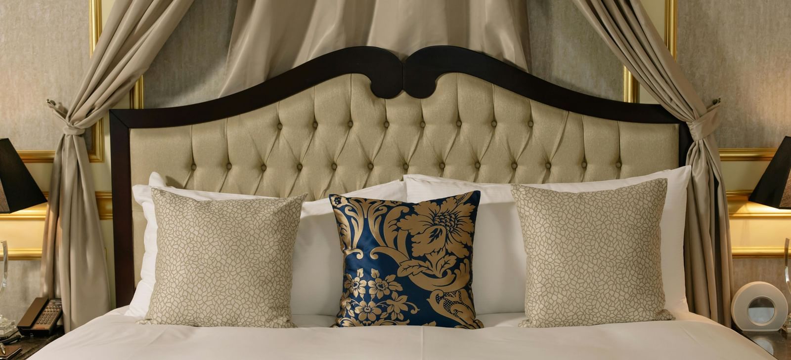 A luxurious hotel room features a large bed with a canopy and elegant bedding, flanked by bedside tables with lamps.