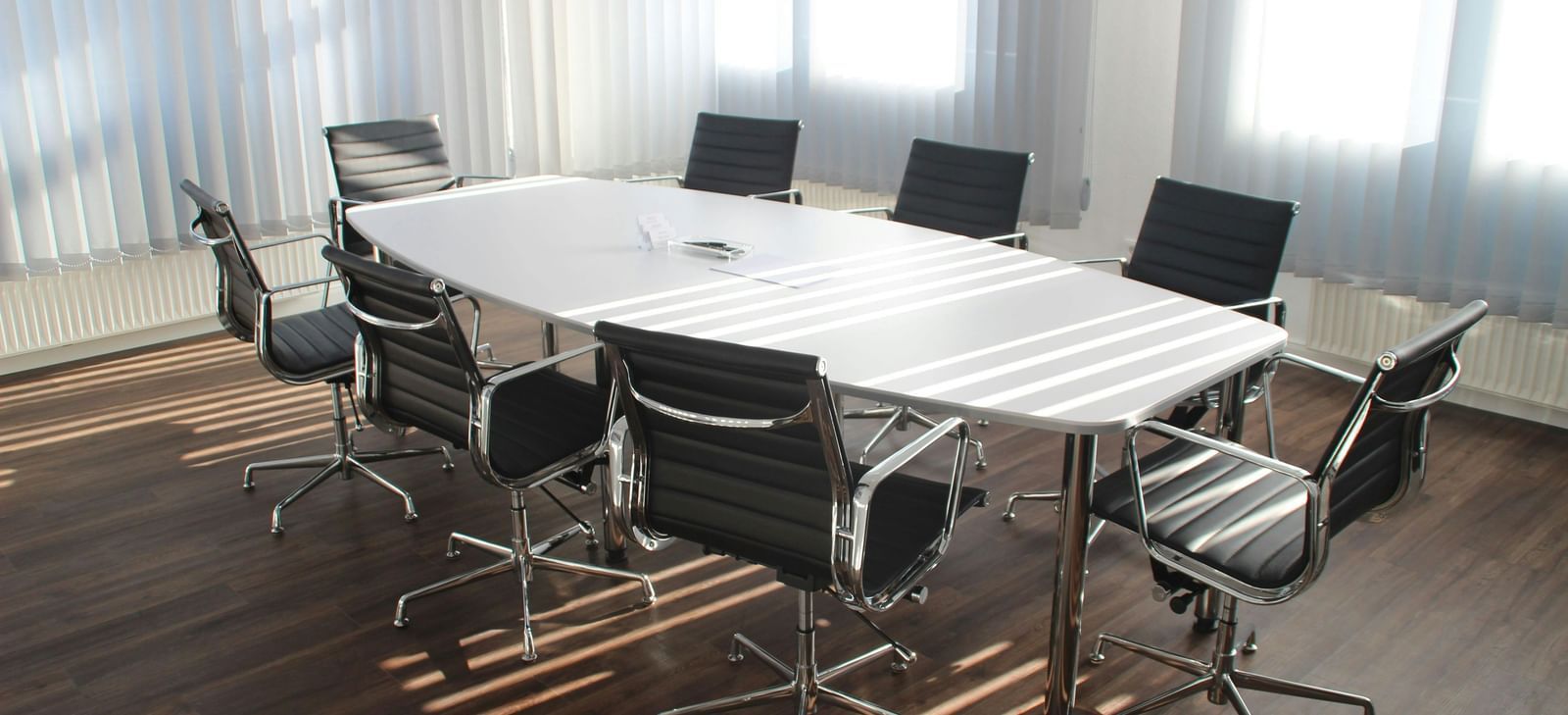 A conference room with a large oval table surrounded by office chairs, illuminated by natural light from windows.