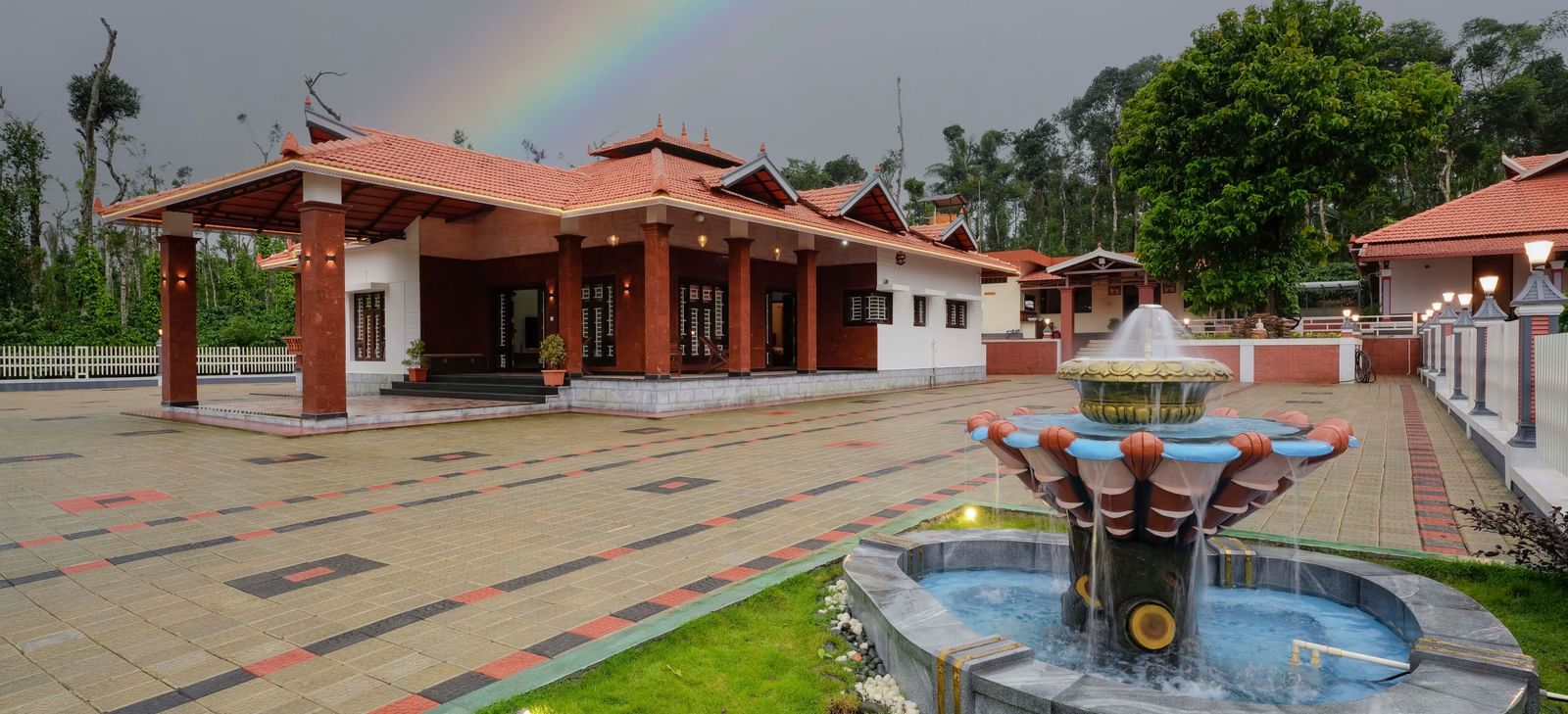 The traditional facade of Bhuthanakadu Retreat under a vibrant rainbow, featuring a fountain in a garden and a colorful patterned walkway leading to the main buildings.