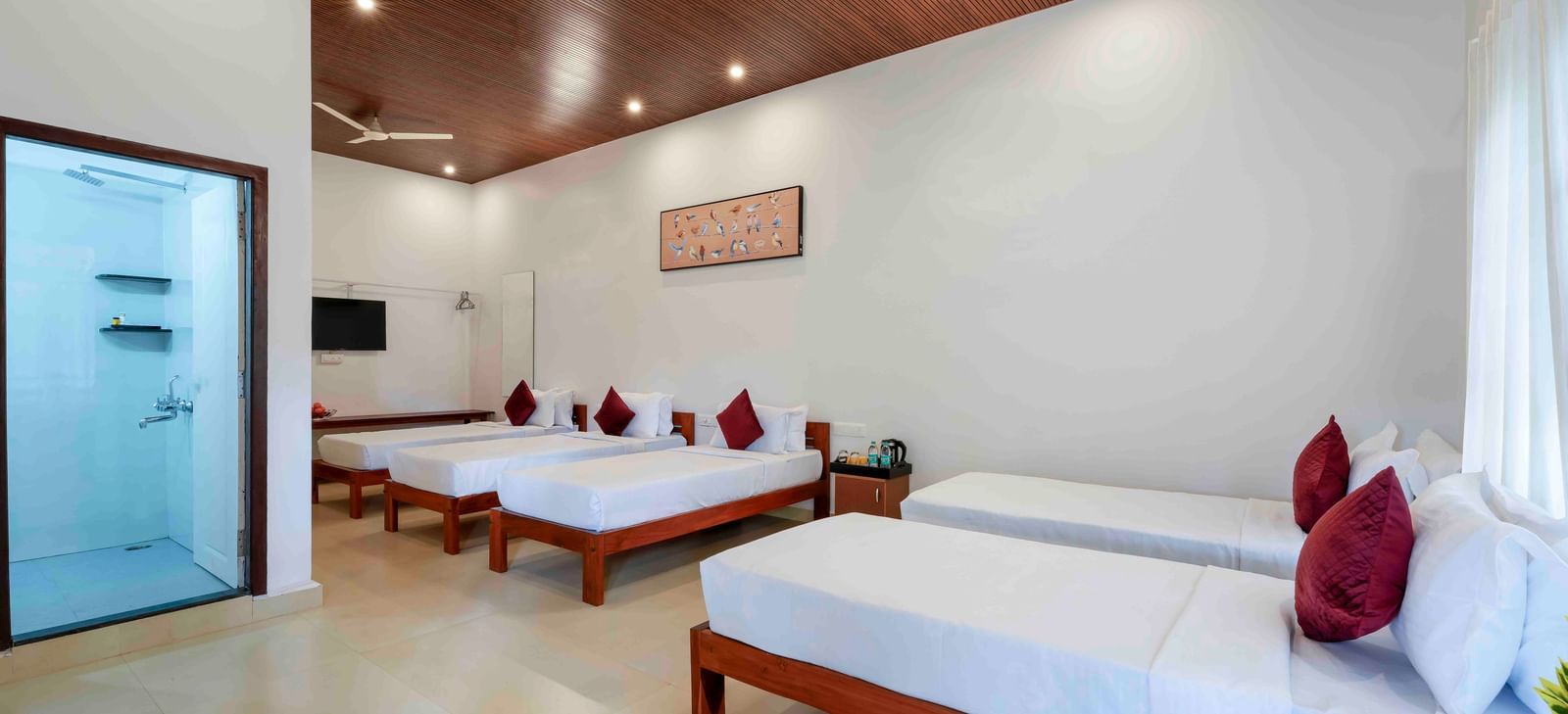 A spacious hall of residences at Bhuthanakadu Retreat, showcasing four single beds with white bedding and red pillows, a modern bathroom, and a warm wooden-paneled ceiling.
