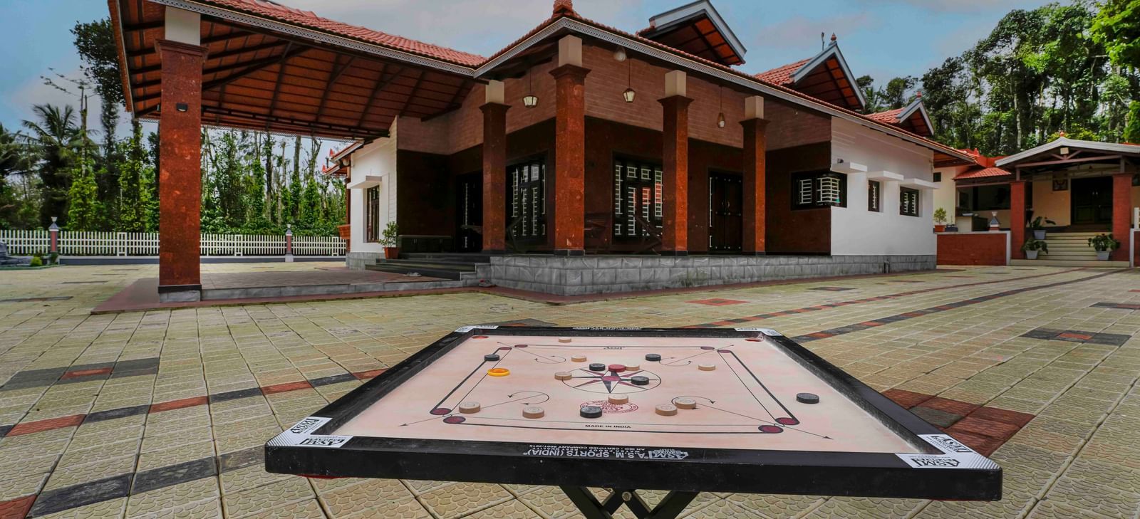 A carrom board is set up outdoors on a paved courtyard, with the traditional architecture of Bhuthanakadu Retreat visible in the background.